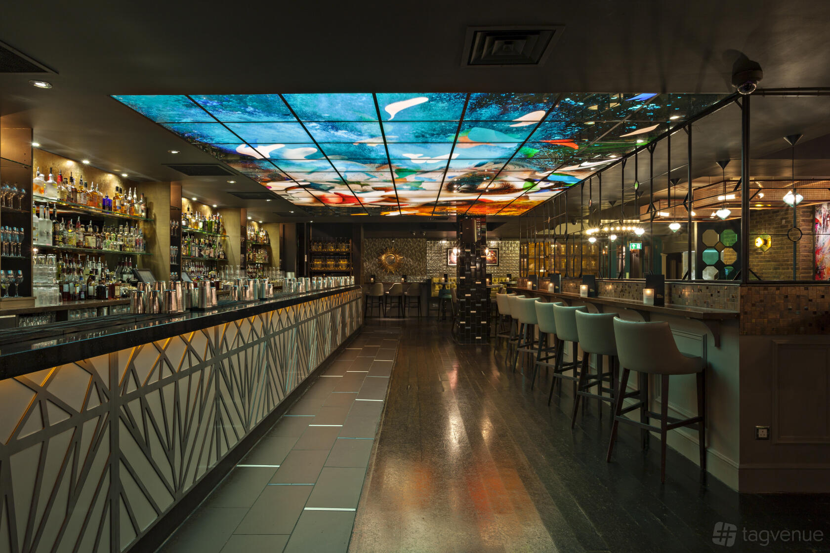 A cocktail bar with an illuminated ceiling display, long mirrored bar, and high-backed stools at Dirty Martini Bishopsgate.