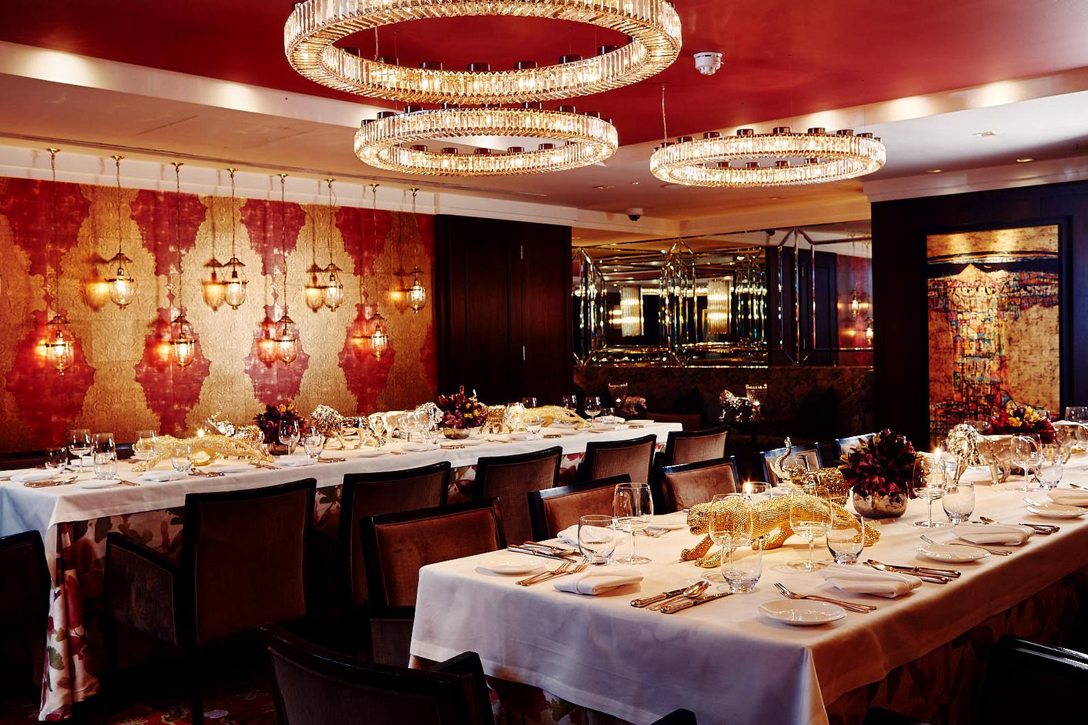 A dining room with ornate chandeliers, gold-patterned wallpaper, and banquet tables with white linens at Chutney Mary.