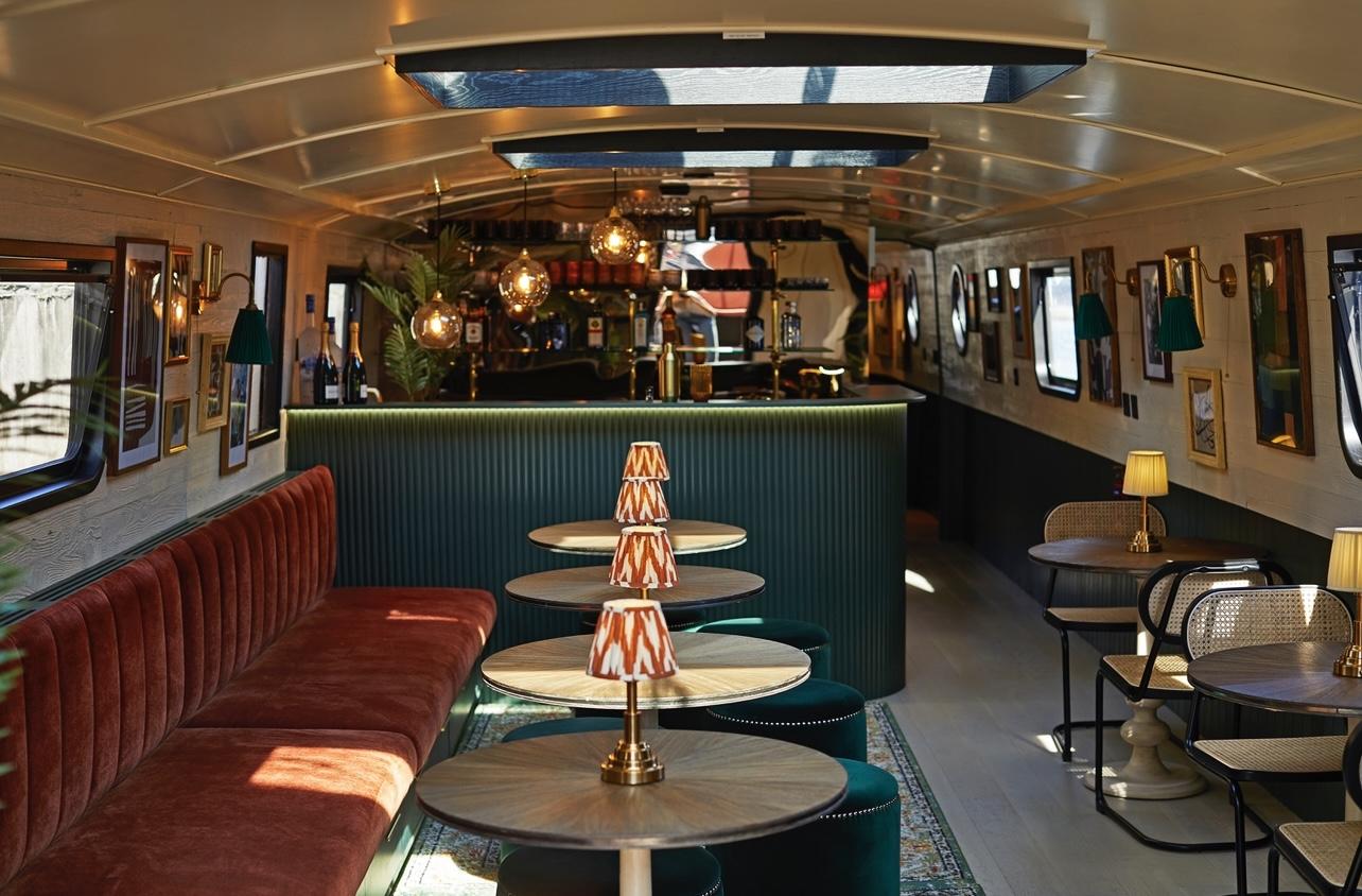 A bar on a boat with velvet banquette seating, round tables, patterned lamps, and wall art at The Boathouse London - Canary Wharf.