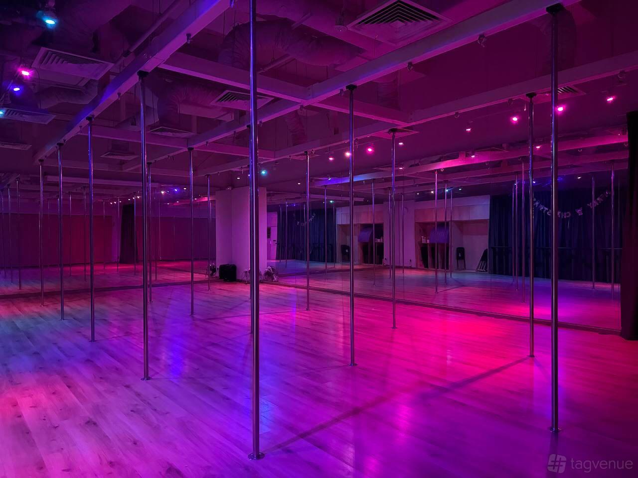 A dance studio with mirrored walls, hardwood floors, and ceiling-mounted poles under purple lighting at Diva Pole Academy.