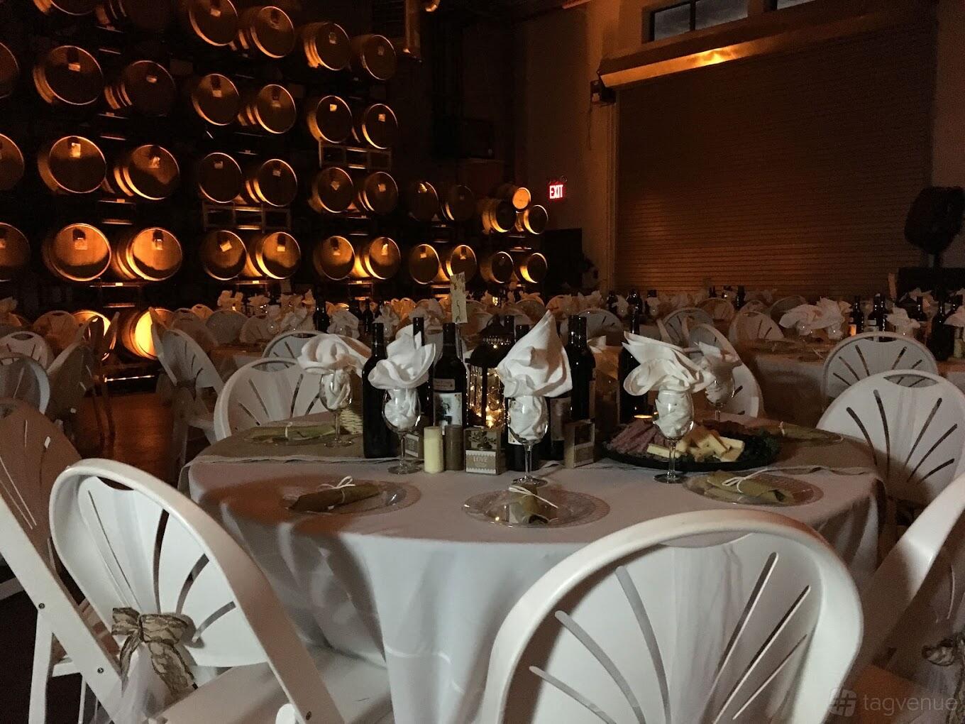 A restaurant with round tables set with white linens and napkins beside a wall of wine barrels at Vino Divino Winery.