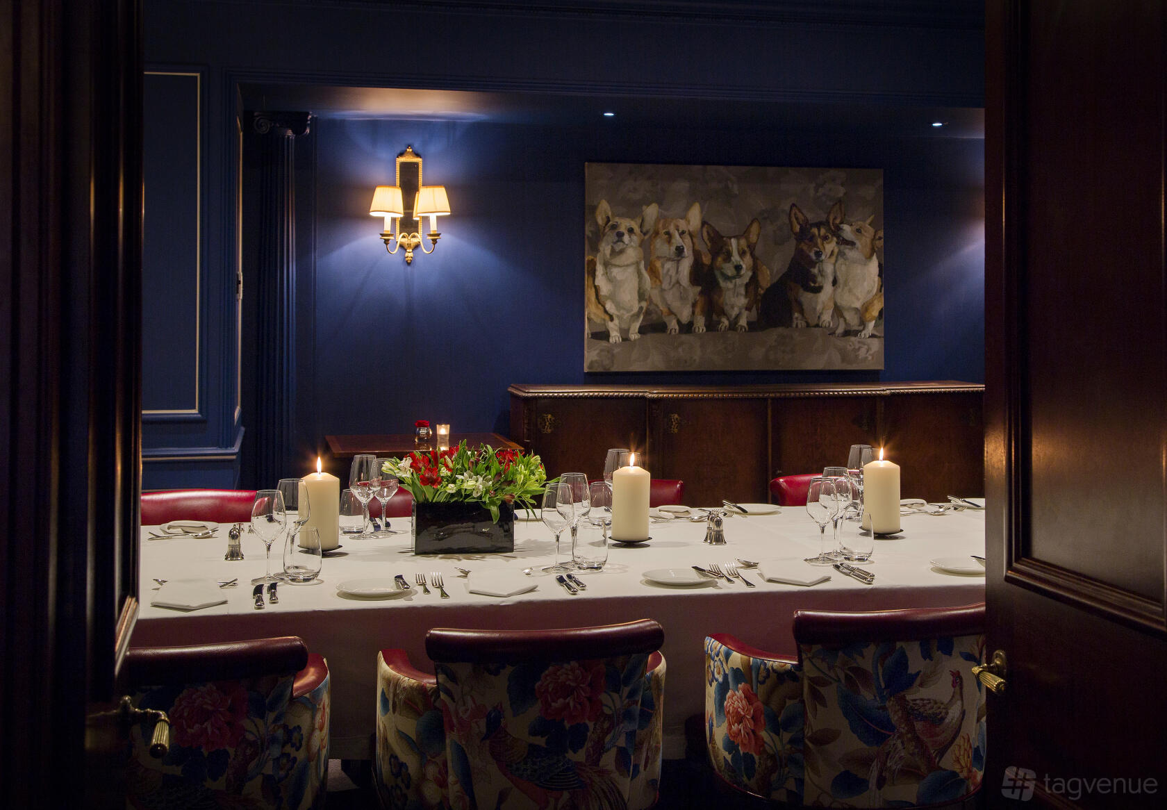 An elegant dining room with a long table, red upholstered chairs, candles, and corgi dog artwork at The Stafford London.