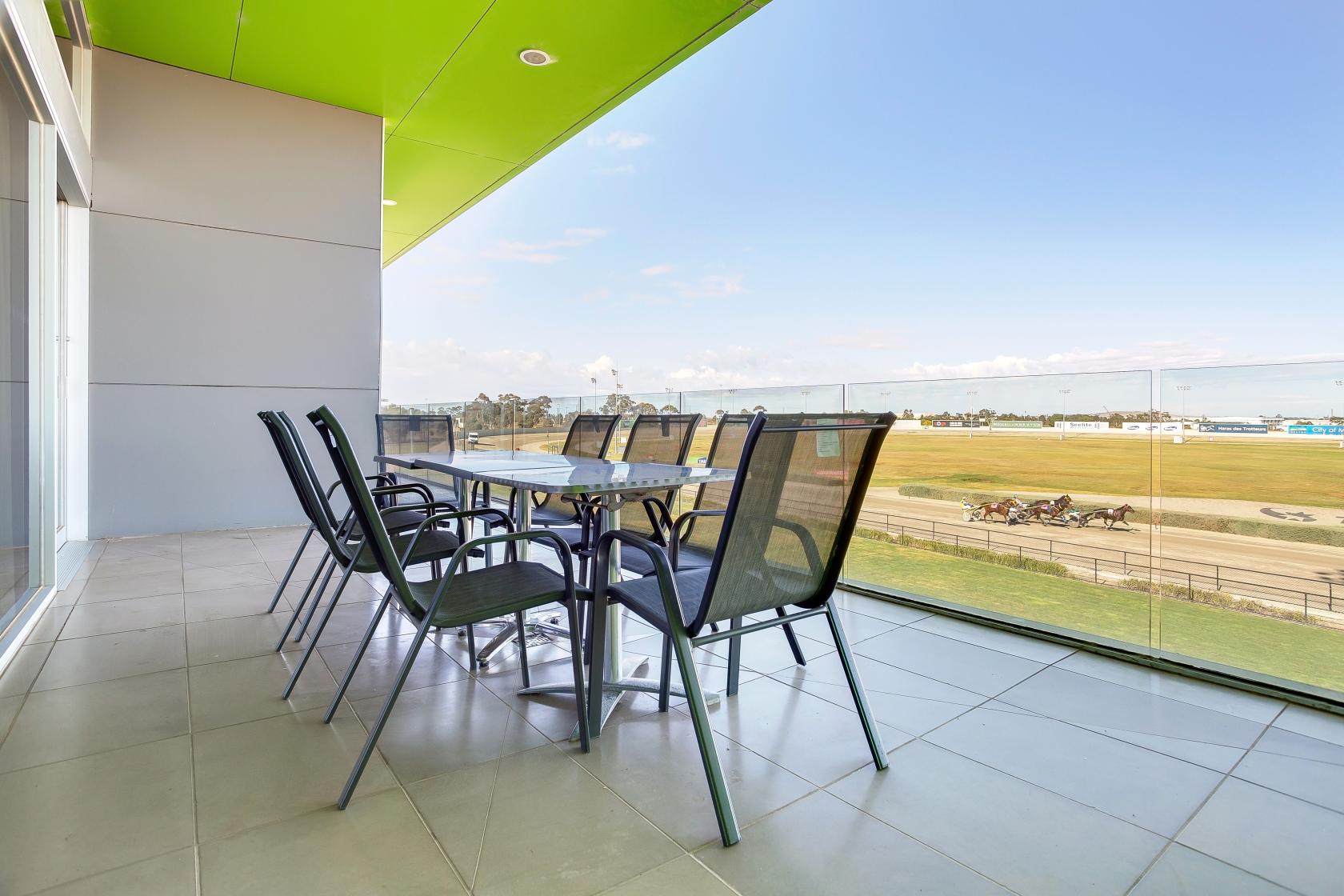 An outdoor balcony with a glass railing, long table, and mesh chairs overlooking a racetrack at Melton Entertainment Park.