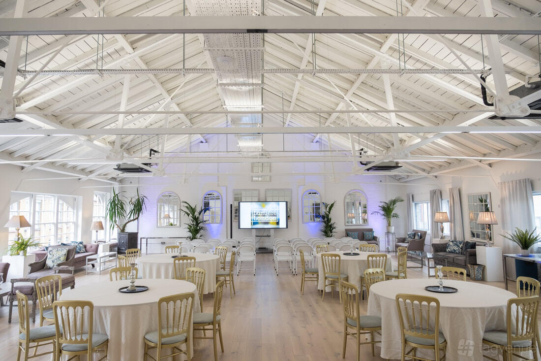 A loft event space with white-painted exposed beams, round tables, and large windows at Lumiere London Old Street.