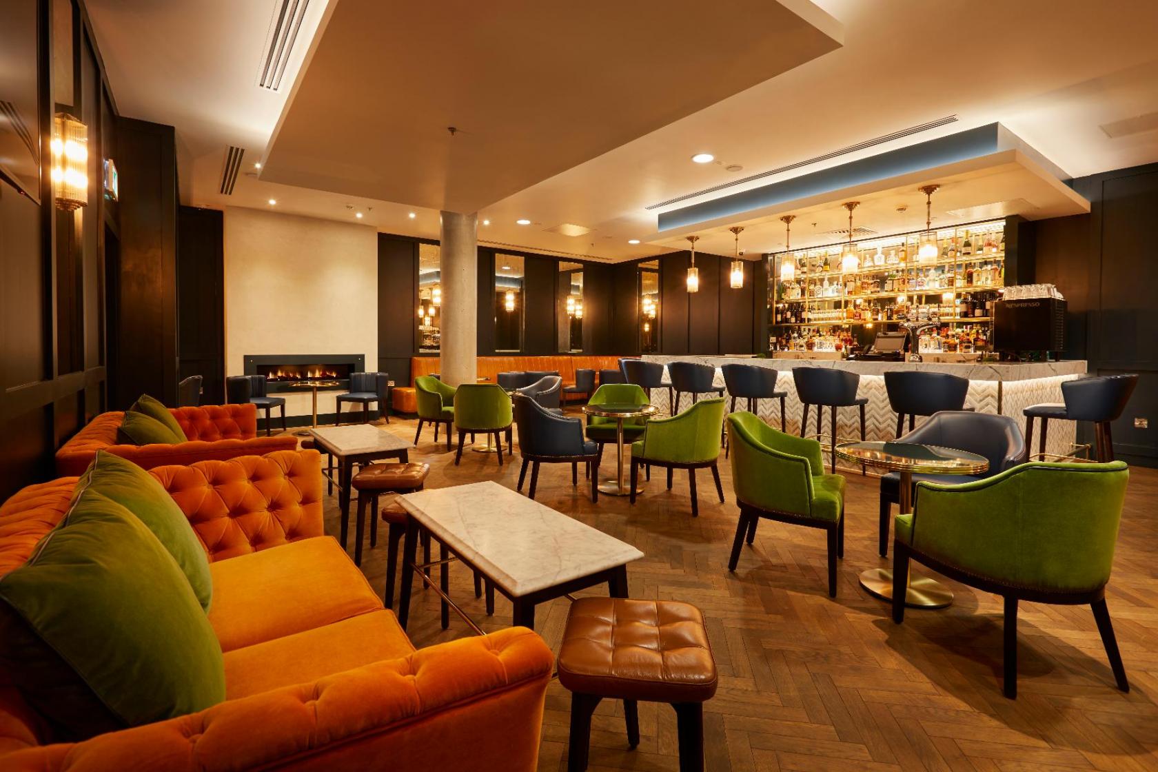 A hotel bar with colorful lounge chairs, velvet sofas, marble tables, and a backlit bar counter at Marlin Waterloo.