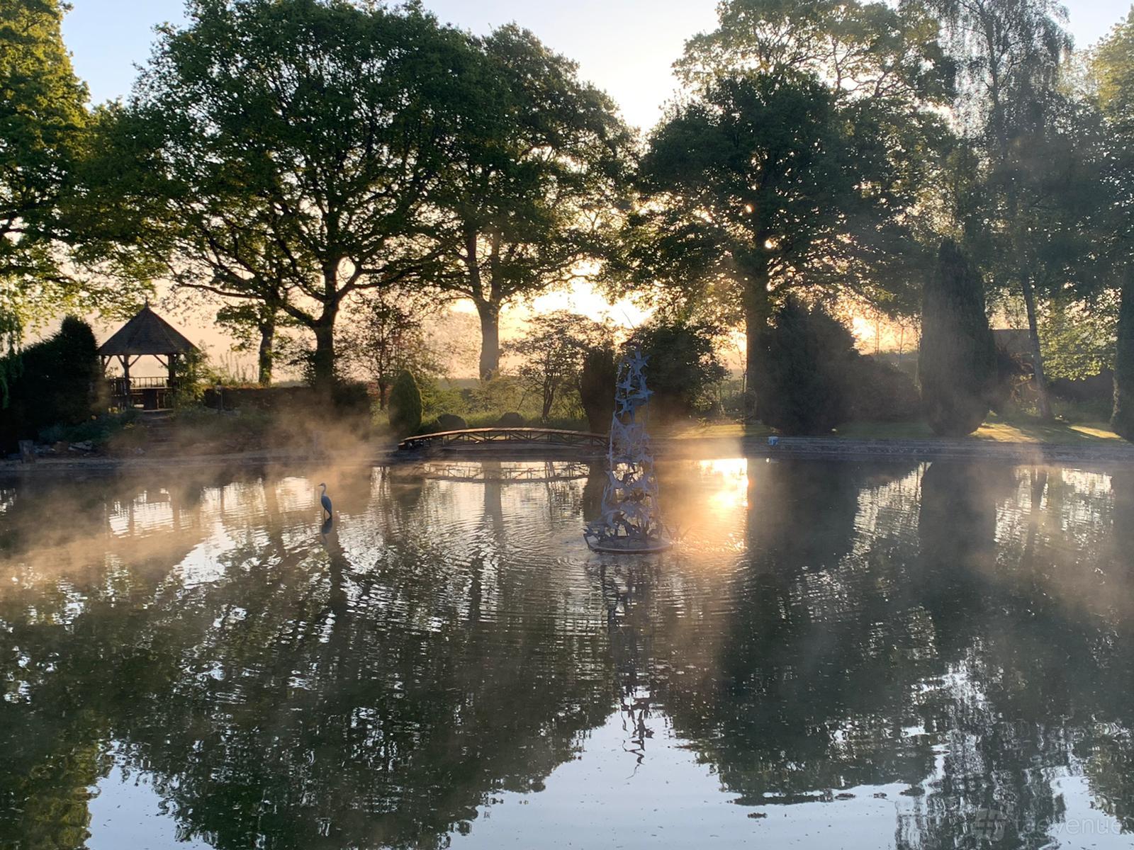 A country house garden with a misty pond, central fountain sculpture, and mature trees at Bullard's Farm.