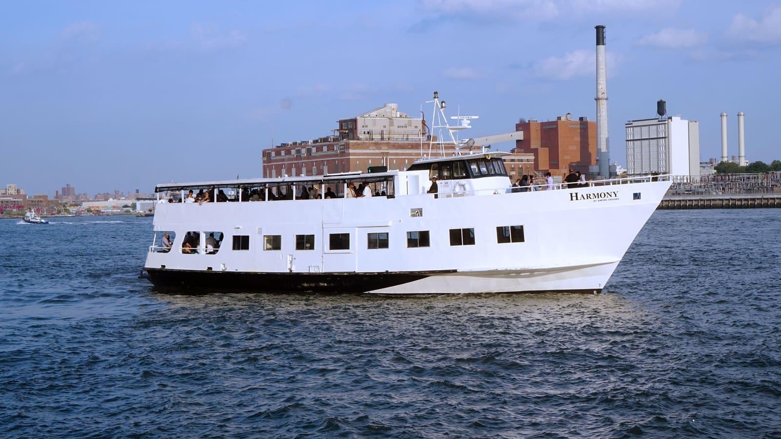A white charter boat with an upper open-air deck cruising on the river near industrial buildings at Empire Cruises.