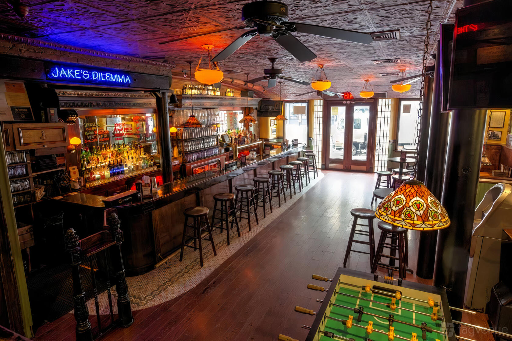 A bar with a long wooden counter, vintage ceiling tiles, amber pendant lighting, and a foosball table at Jake's Dilemma.
