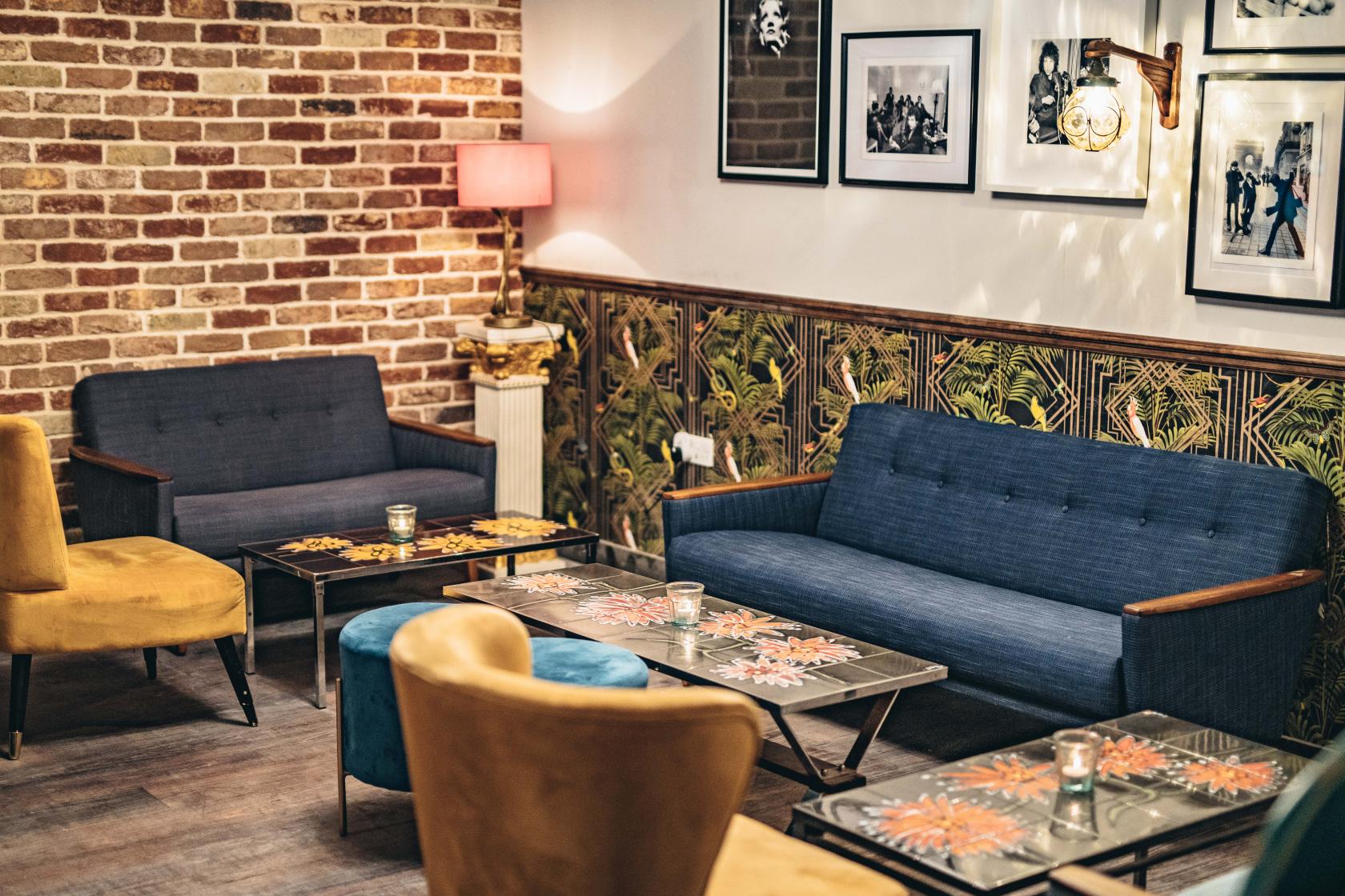 A lounge in a cocktail bar with exposed brick walls, blue sofas, yellow chairs, and patterned wallpaper at Callooh Callay Chelsea.