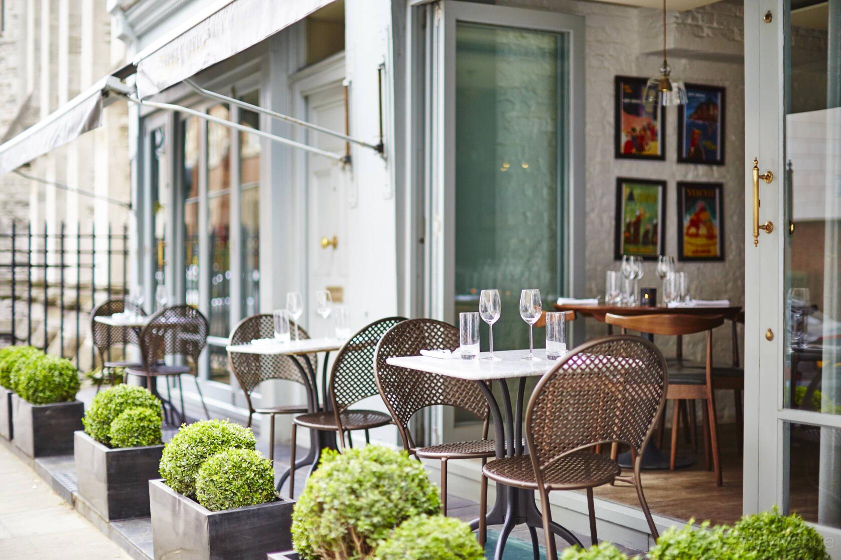 An outdoor restaurant space with round tables, woven chairs, and potted greenery at Trishna.