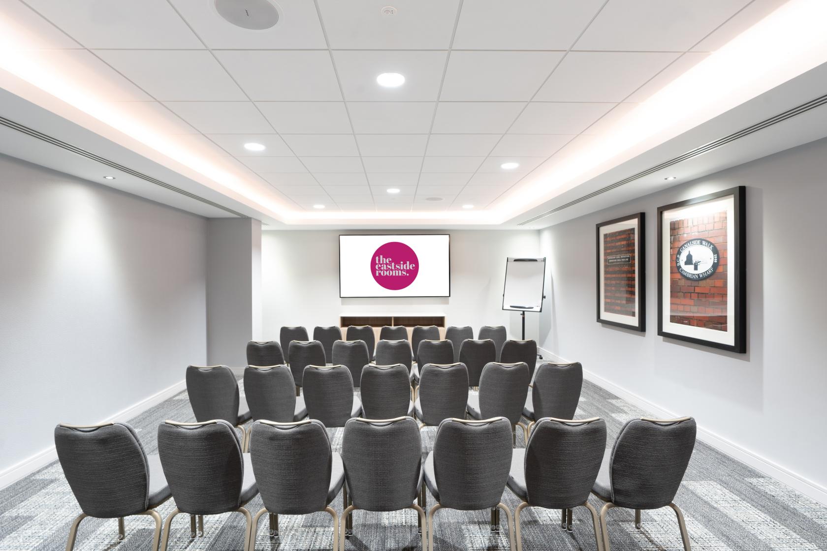 A meeting room with rows of grey chairs, a large presentation screen, and framed artwork at The Eastside Rooms.