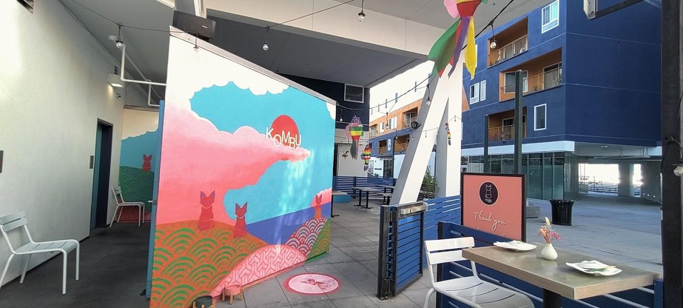 An outdoor patio with colorful mural, blue bench seating, string lights, and shaded tables at Kombu Sushi Arts District.