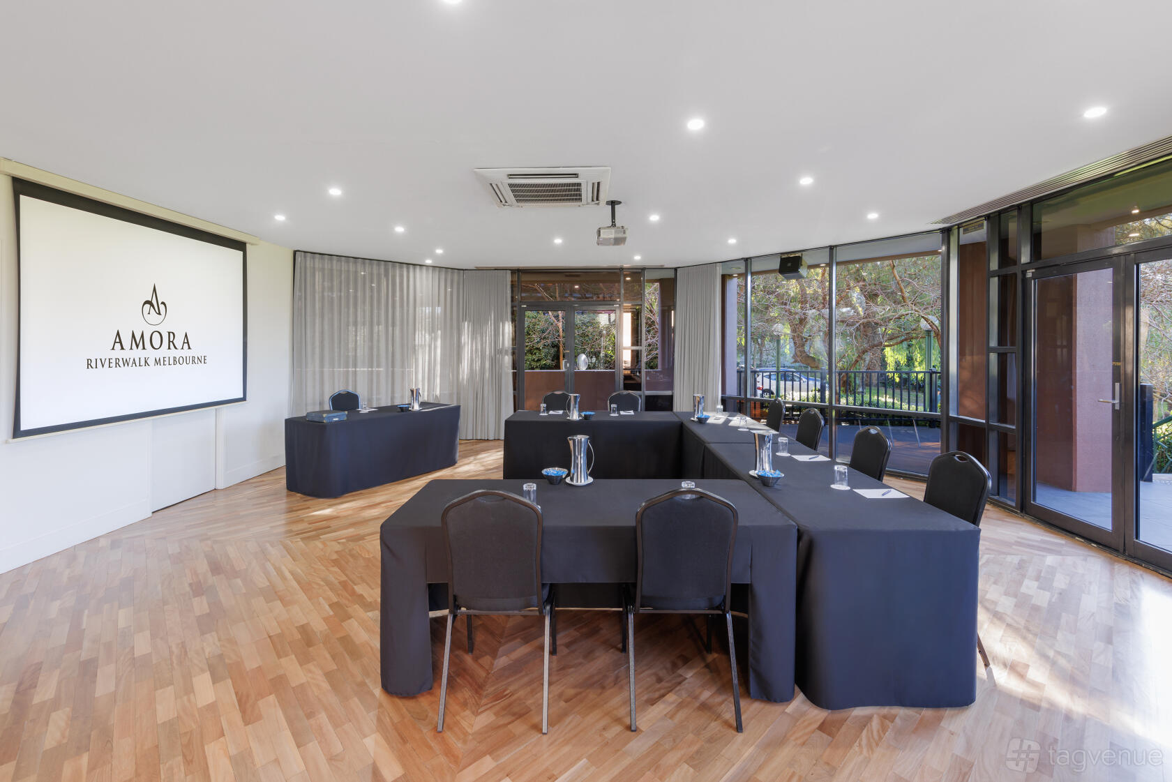 A function room with parquet flooring, natural light from floor-to-ceiling windows, and U-shaped tables at Amora Hotel Riverwalk Melbourne.