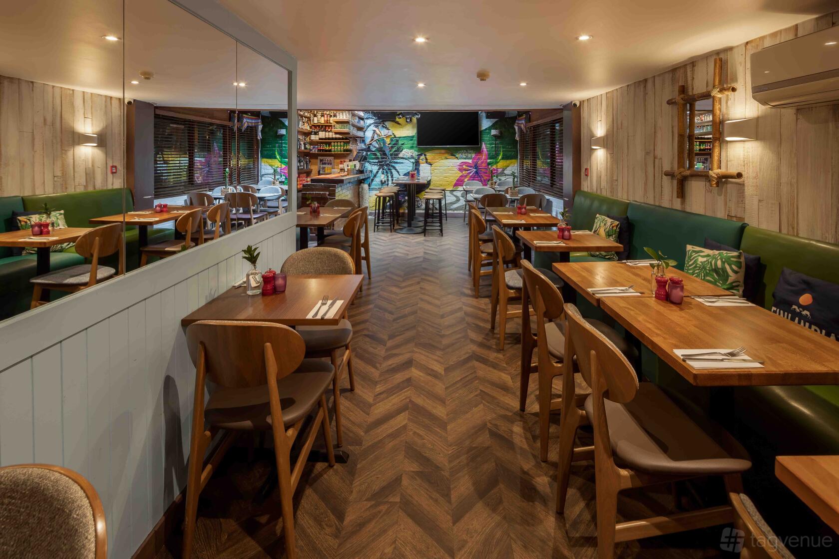 An event space in a restaurant with wooden tables, green banquette seating, and tropical mural at Made in Brasil - Camden Town.