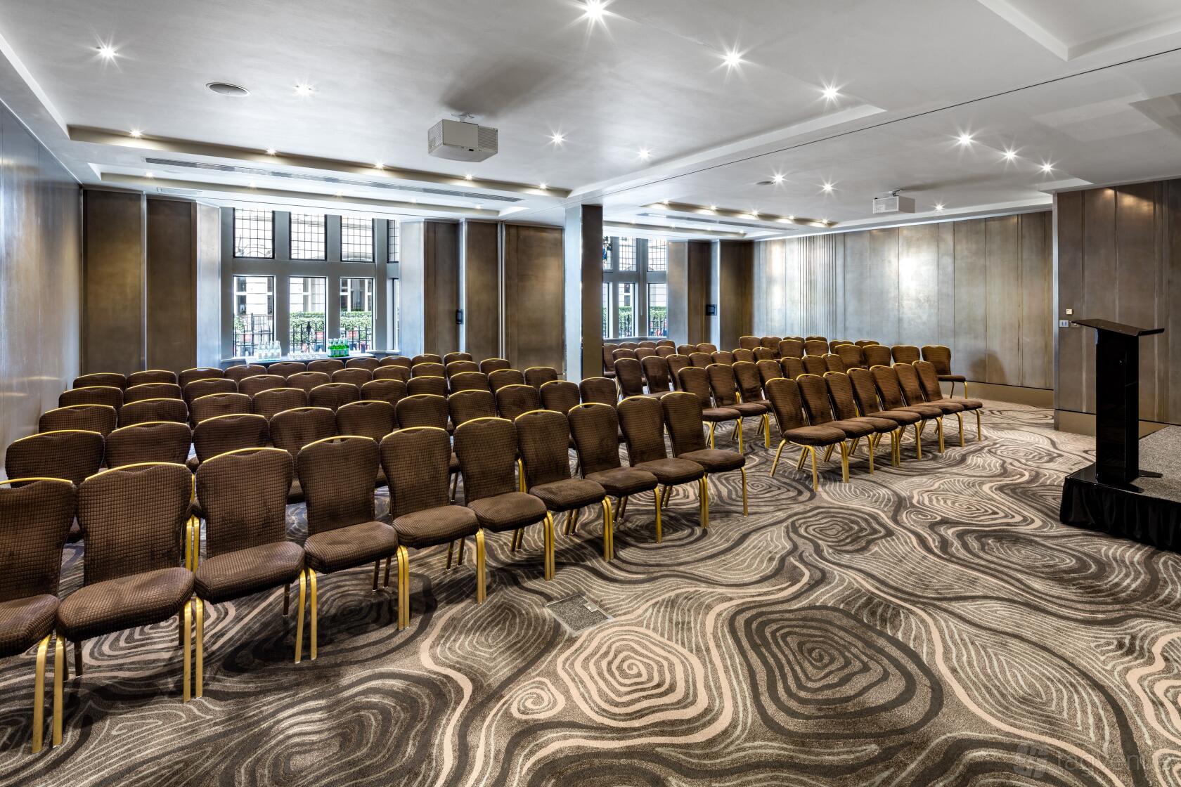 A meeting room with rows of brown cushioned chairs, patterned carpet, and floor-to-ceiling windows at Radisson Blu Hotel, London Bloomsbury.