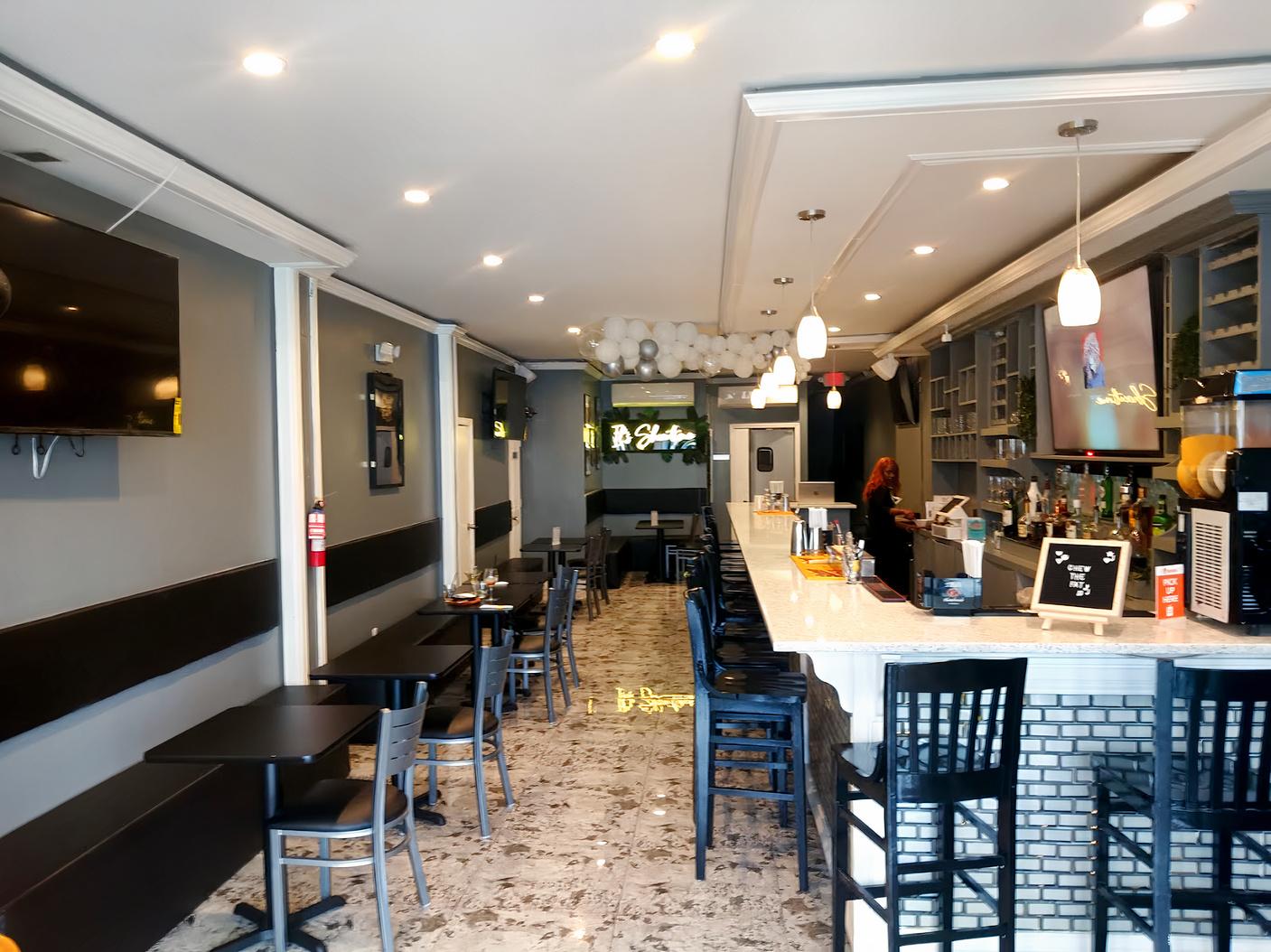 A bar with a long marble counter, pendant lighting, black chairs, and a tiled floor at Chew The Fat Bar, Lounge & Grill.