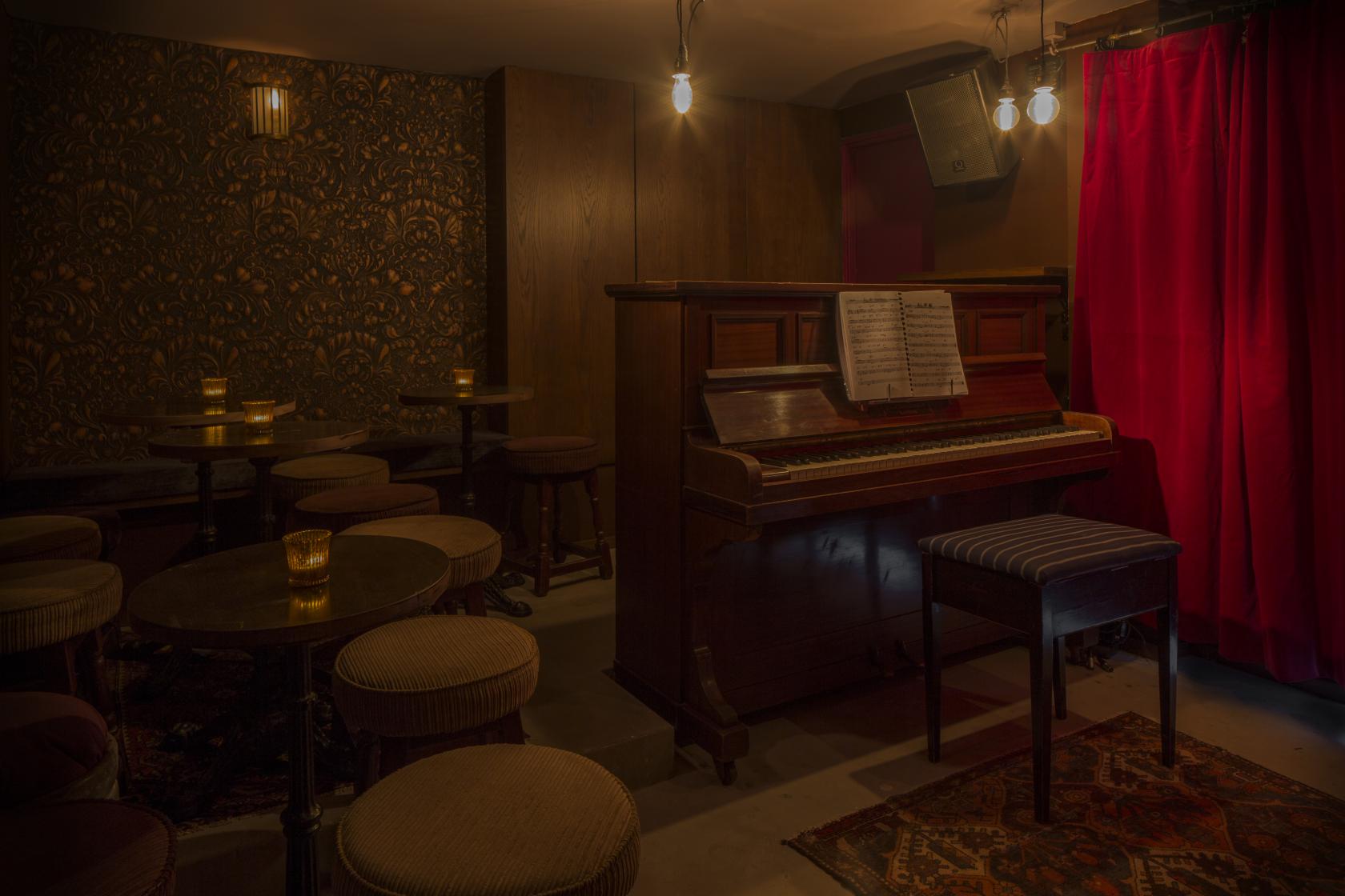 A basement bar with patterned wallpaper, an upright piano, red curtains, and small candlelit tables at Green Note.