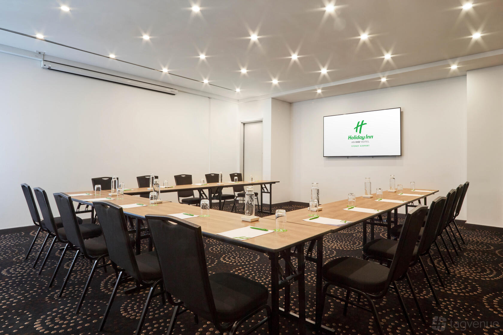 A hotel meeting room with U-shaped tables, black chairs, and a large wall-mounted screen at Crowne Plaza Sydney Airport.