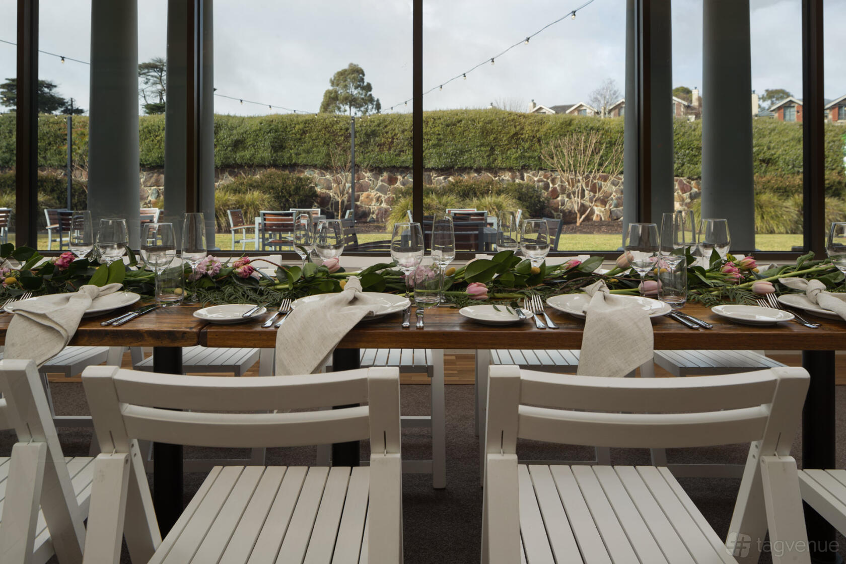 An event space with floor-to-ceiling windows, white chairs, and a long table set with floral arrangements at Yarra Valley Lodge.