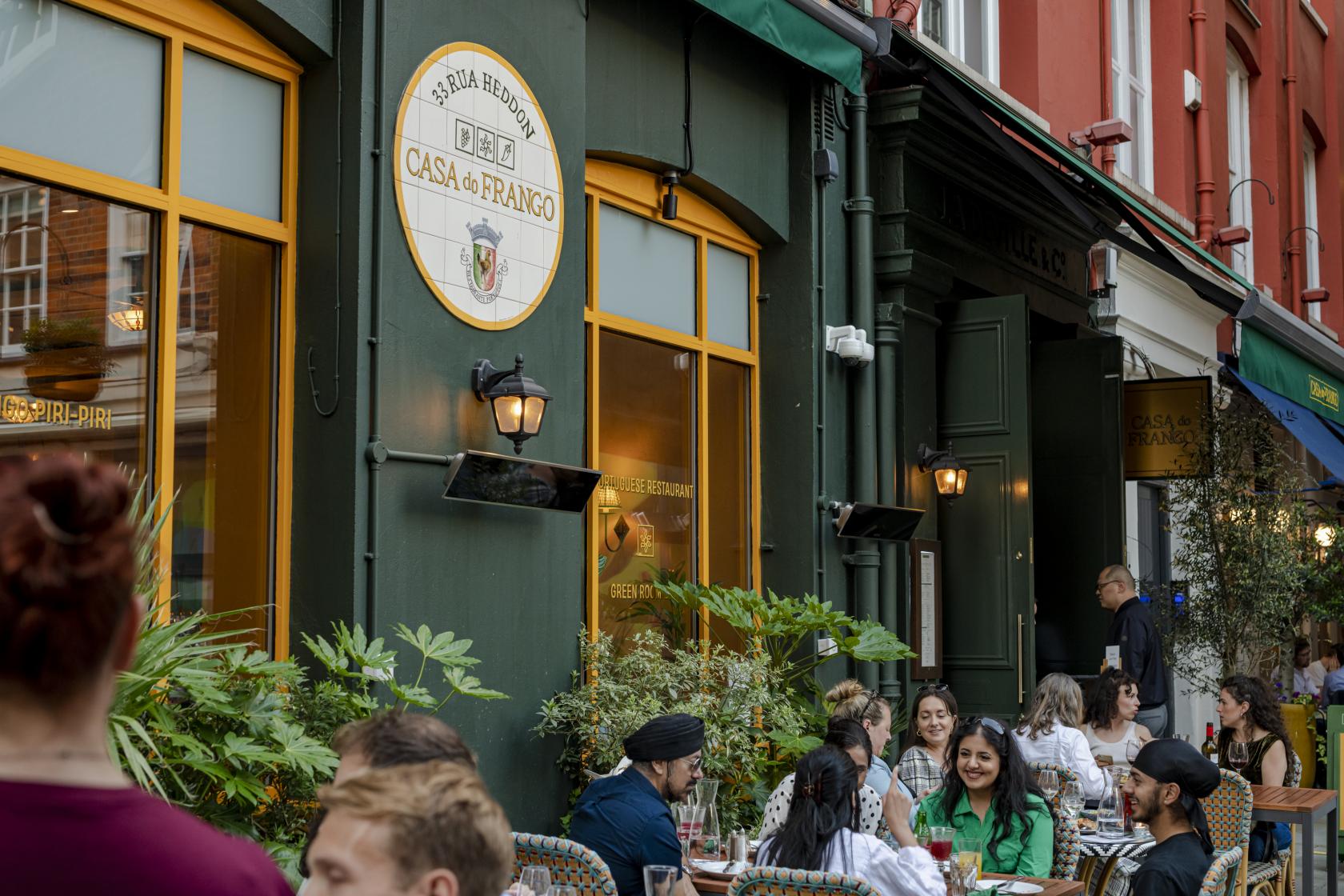 A restaurant with outdoor seating, green façade, large windows, and potted plants at Casa do Frango - Mayfair.