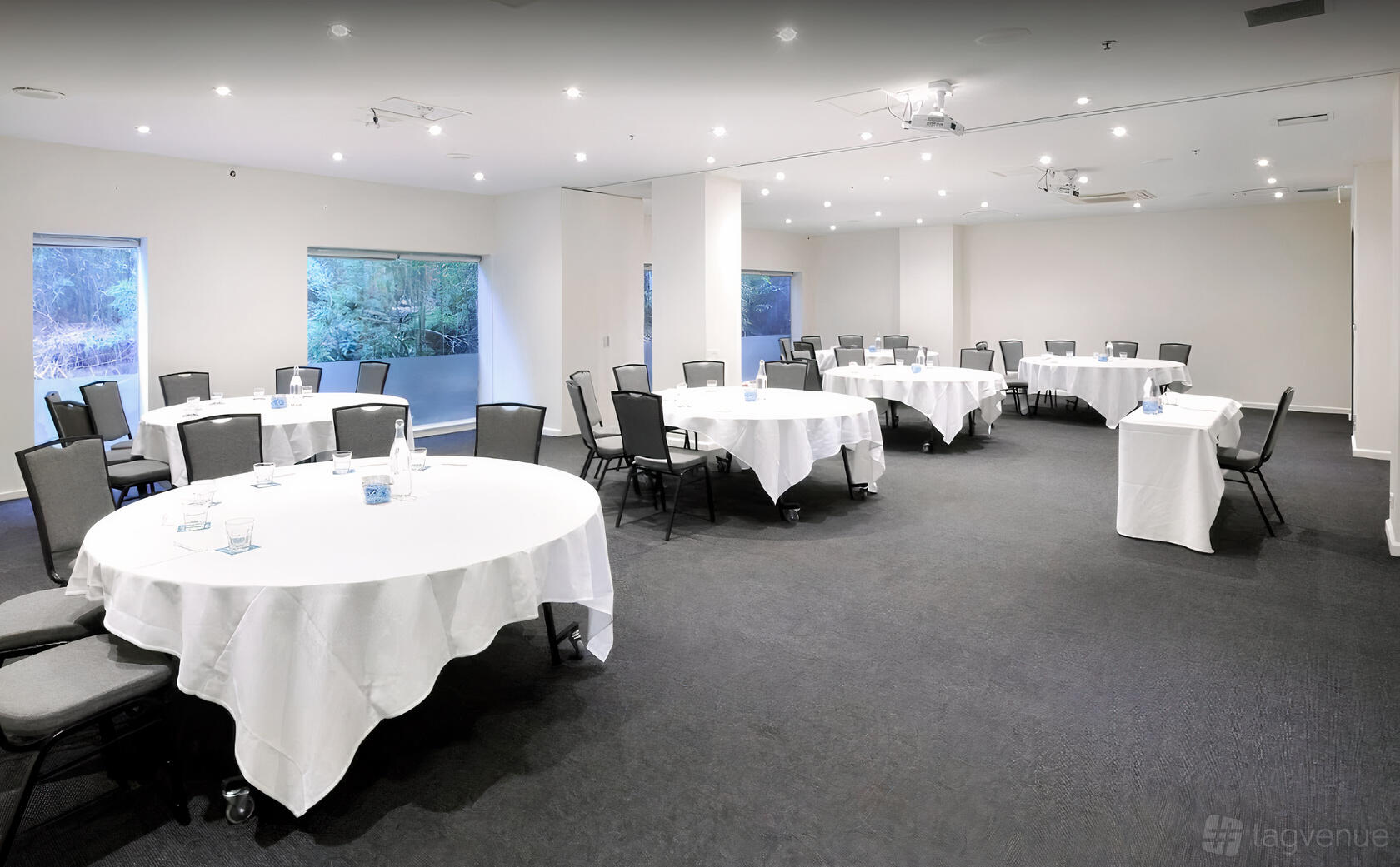 An event space with round tables draped in white linens, black chairs, and natural light at Mercure North Melbourne.