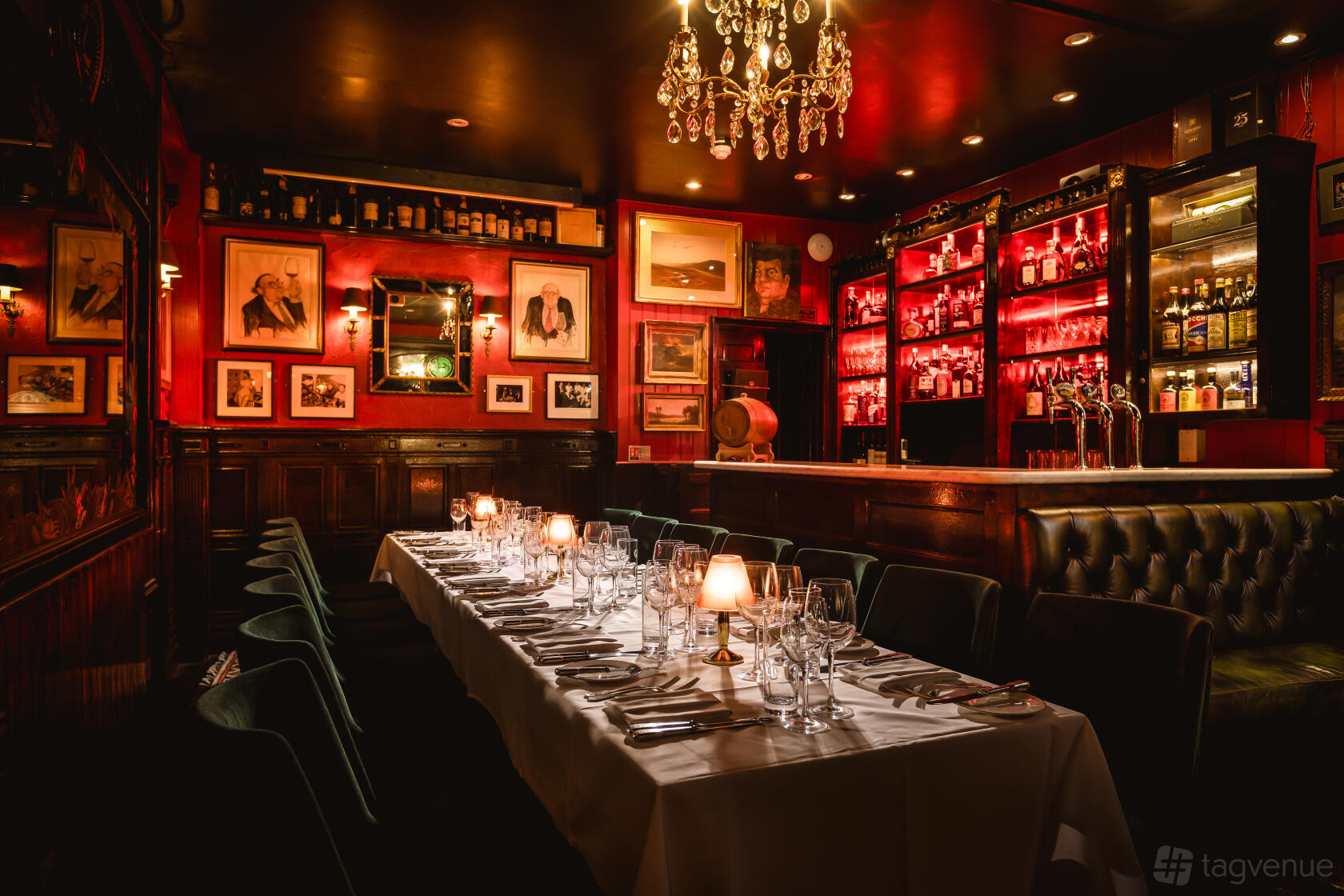 A bar in a restaurant with red walls lined with framed art, chandeliers, and a long table with white linens at Boisdale of Belgravia.