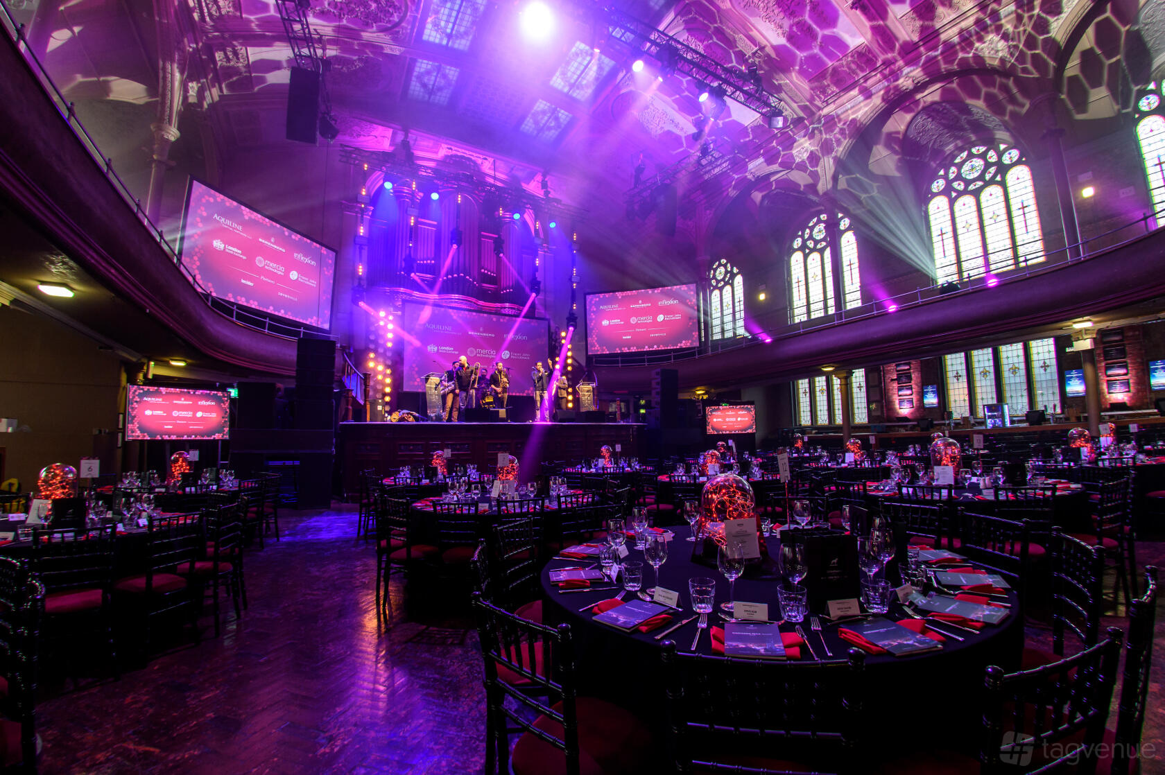 A historic hall with arched stained glass windows, dramatic stage lighting, and round banquet tables at Albert Hall (Manchester).