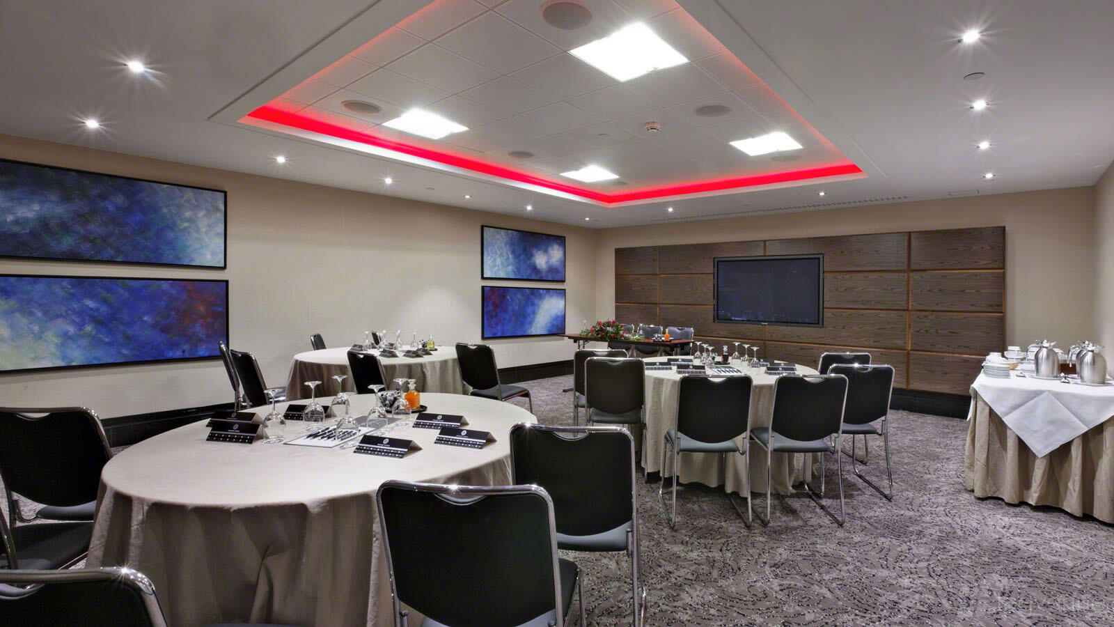 A hotel meeting room with round tables, black chairs, wall artwork, and recessed ceiling lighting at Leonardo Royal London St Paul's.
