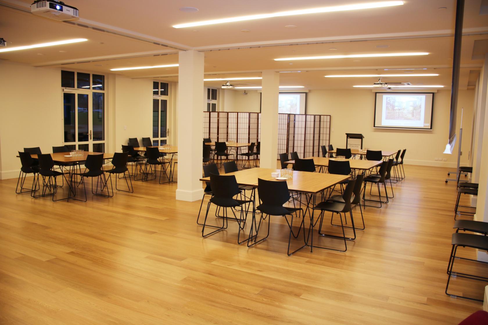 A classroom with wooden floors, movable square tables, black chairs, and ceiling projectors at University College.