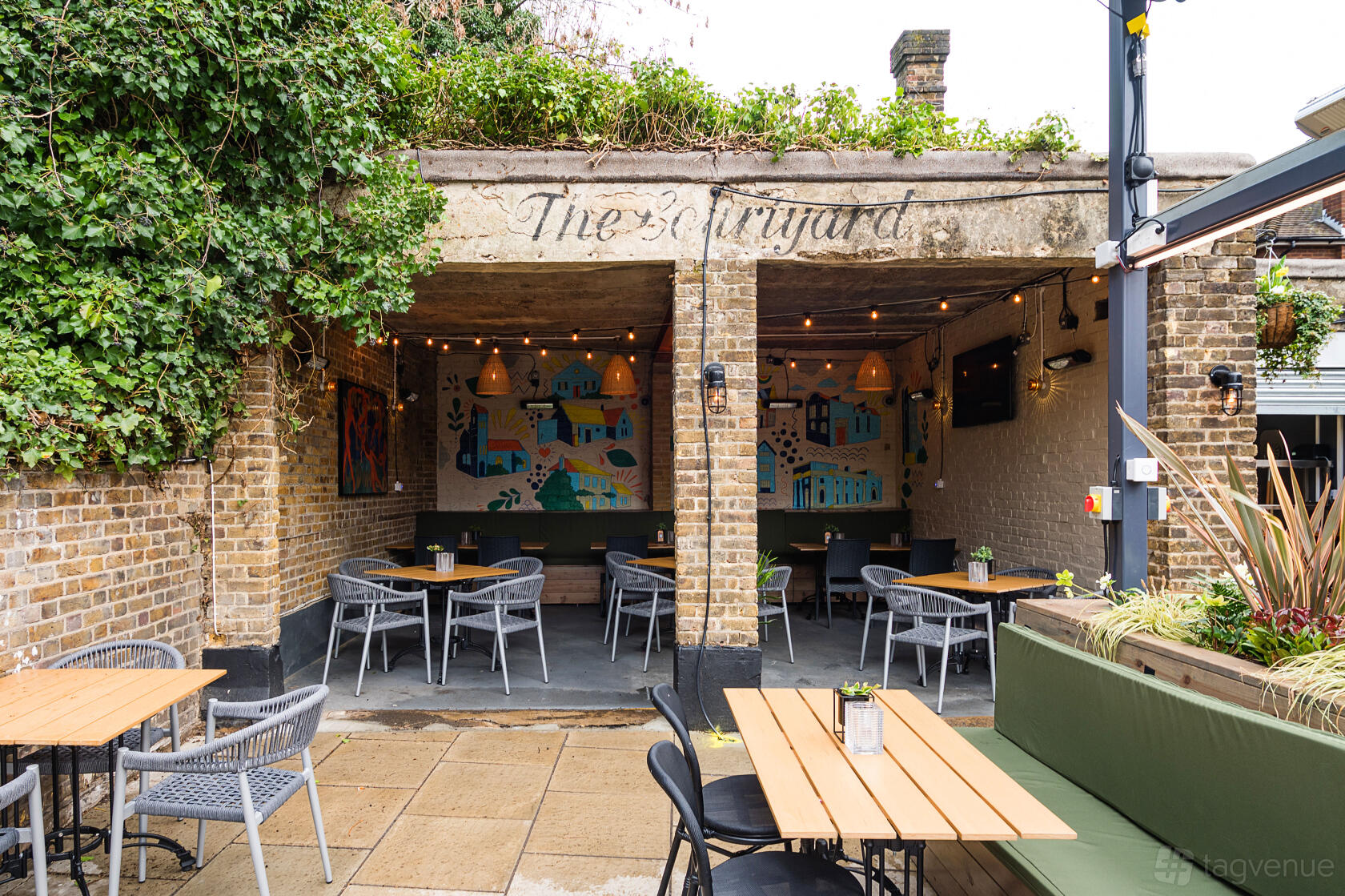 A pub garden with brick walls, wooden tables, grey chairs, string lights, and wall art at The Salisbury Arms Winchmore Hill.