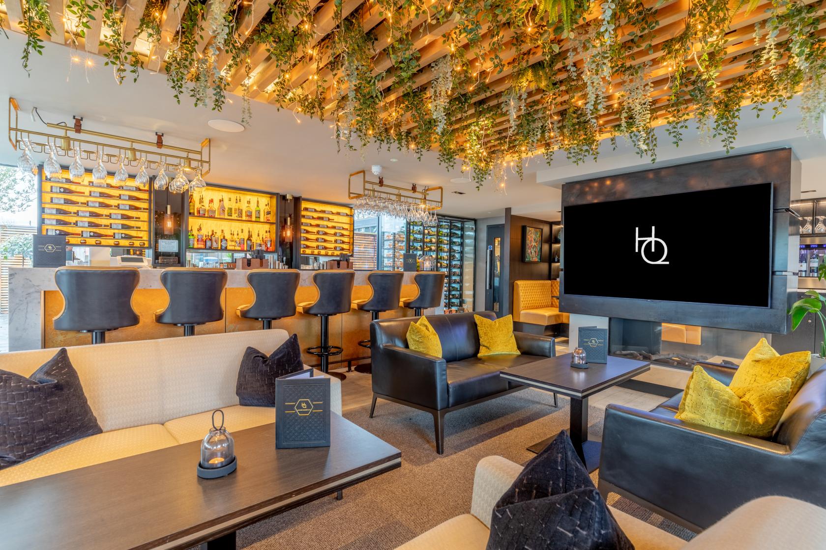 A lounge with a hanging garden ceiling, a stocked bar with stools, and cozy seating at The Colony HQ.