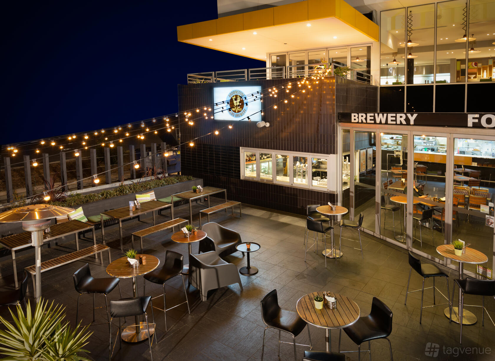 An outdoor bar terrace with string lights, round wooden tables, and metal chairs at Brownstone Micro Brewery.