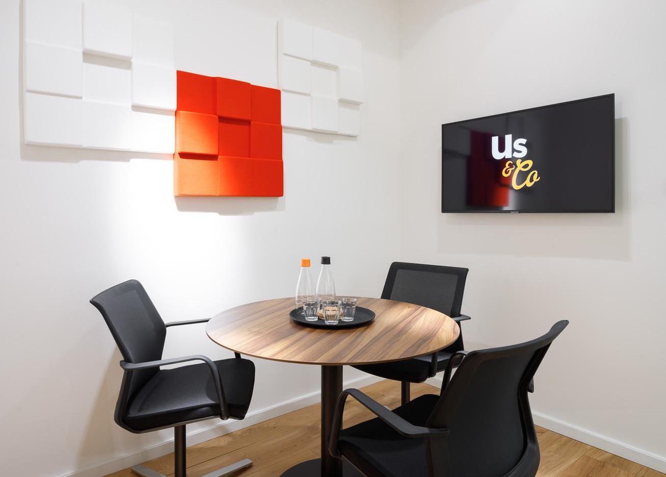 A meeting room with a round wooden table, three black chairs, wall art, and a mounted screen at Us&Co Stratford.