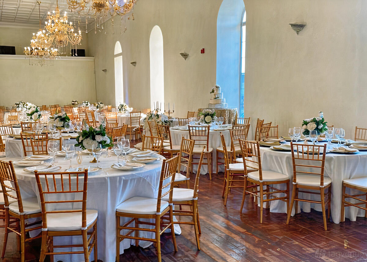 A ballroom with round tables covered in white linens, gold chairs, floral centerpieces, and chandeliers at Elegant Affair Wedding Venue.