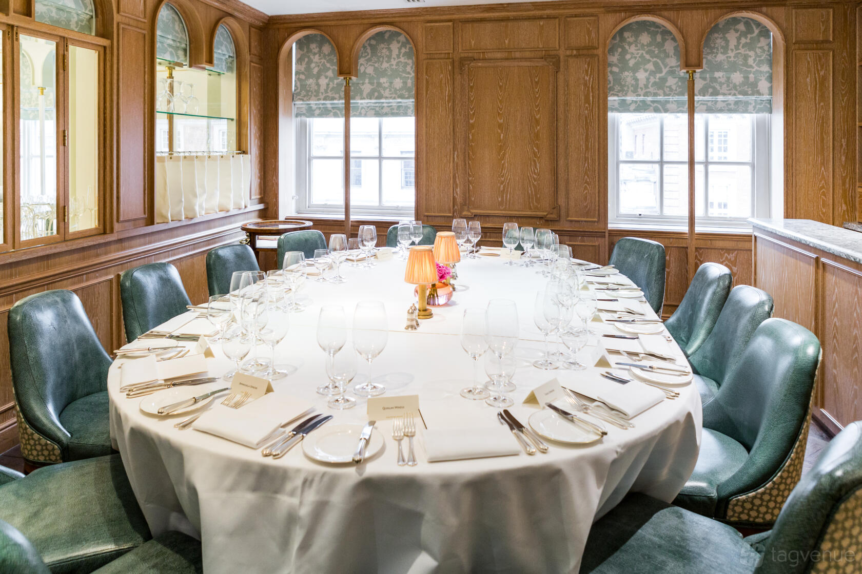 An event venue with an oval table set with glassware and place cards, green leather chairs, and wood-paneled walls at Fortnum & Mason.