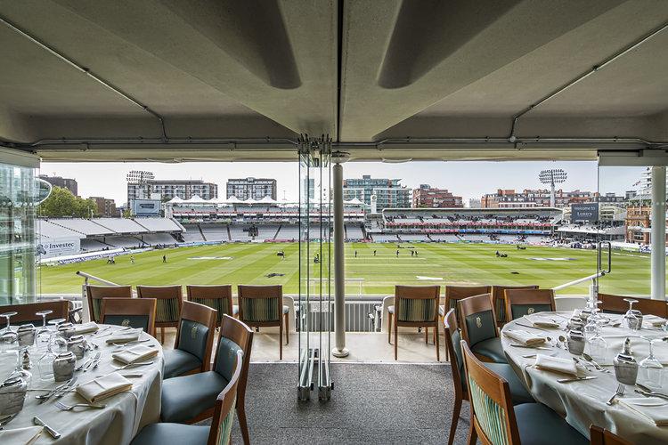 An event venue with round tables set for dining and floor-to-ceiling windows overlooking the cricket field at Lord's Cricket Ground.