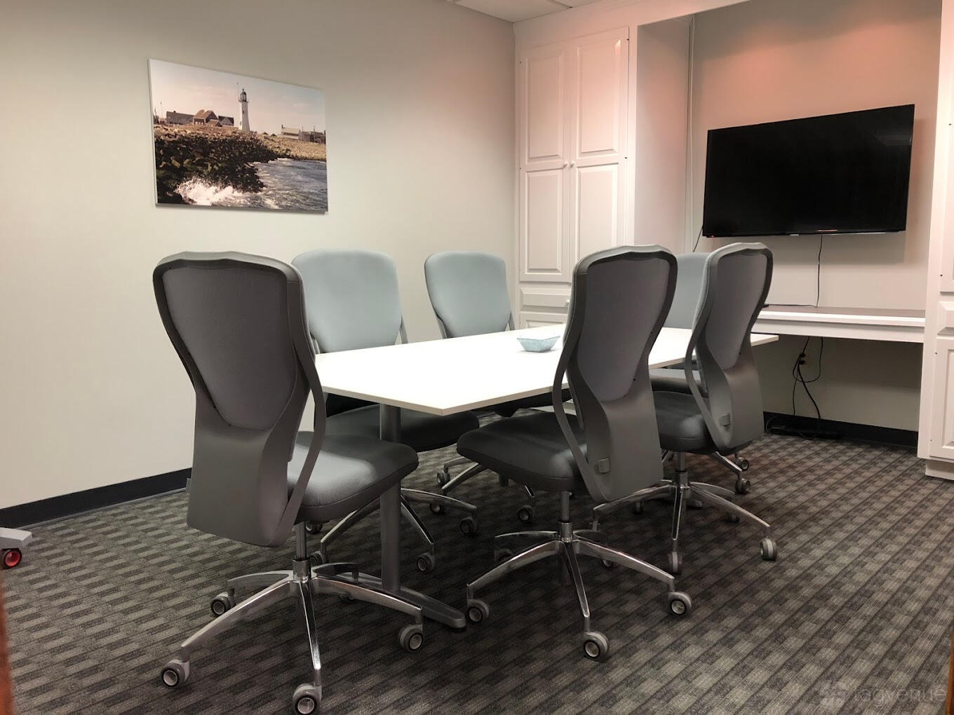 A meeting room with rolling chairs, a white conference table, wall-mounted TV, and lighthouse artwork at Workspace@Shipyard.