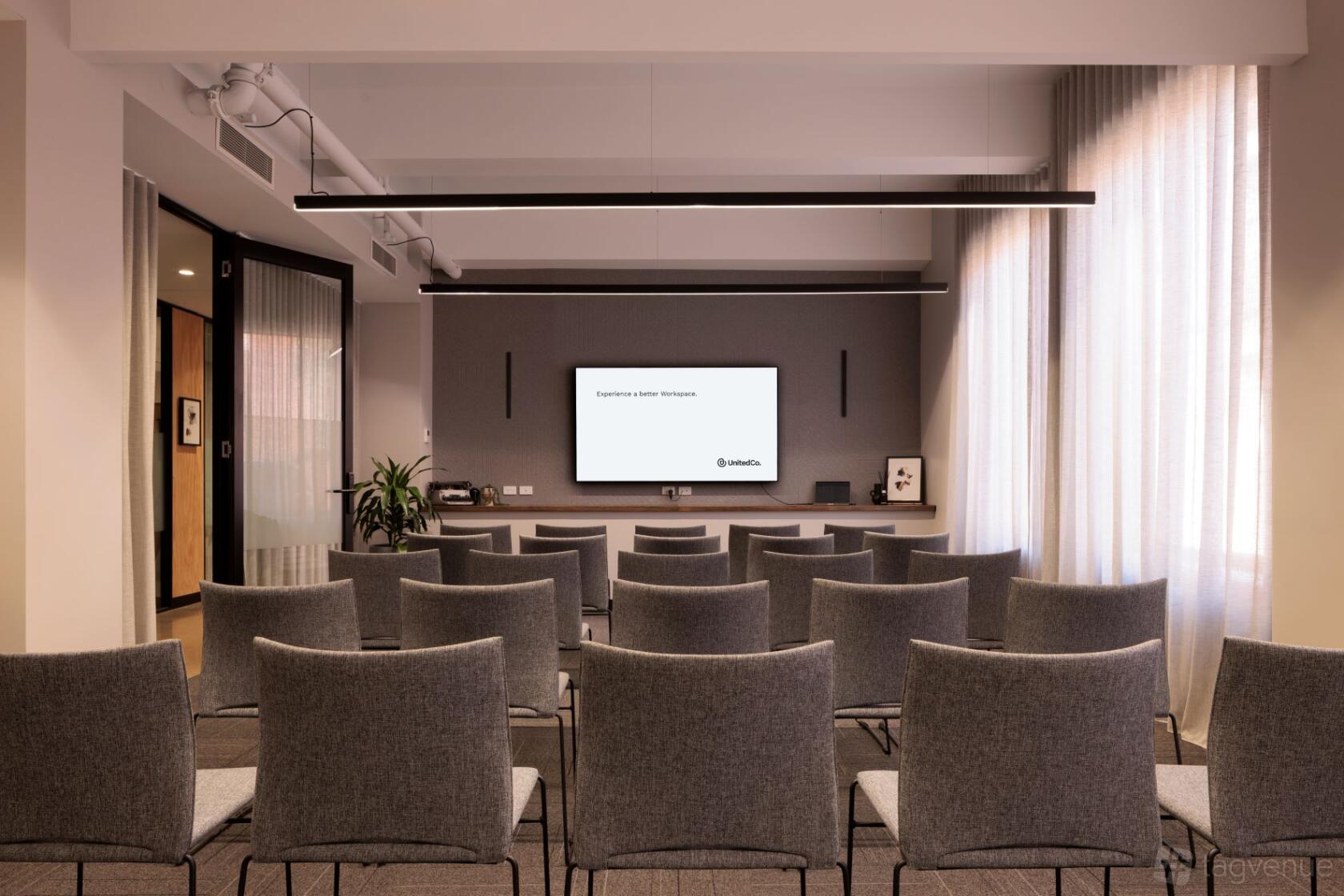 A training room with rows of gray chairs, a large screen, and sheer curtains at United Co. Fitzroy.