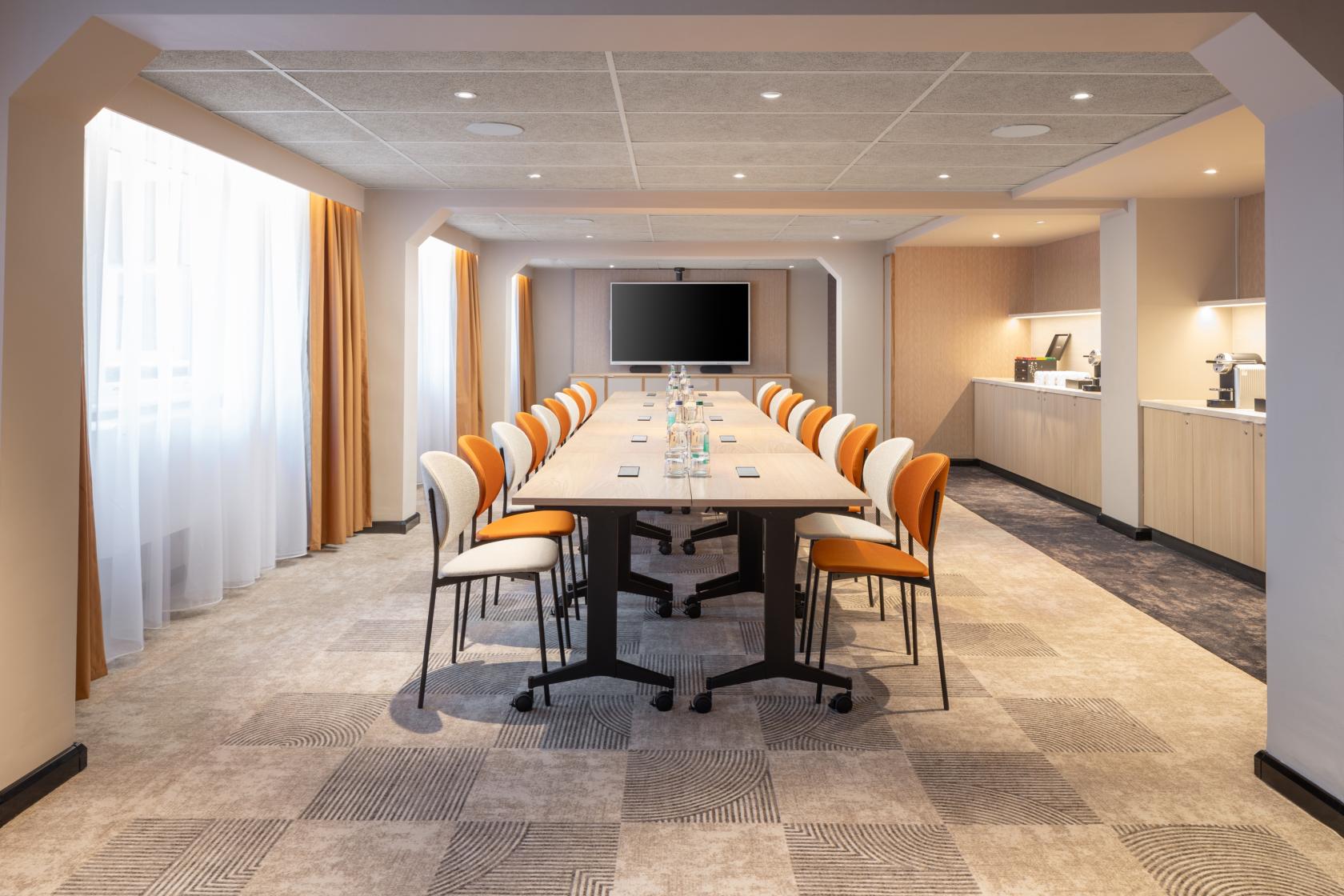 A meeting room with a long conference table, orange chairs, a wall-mounted display, and natural light at Mercure London Earl's Court.