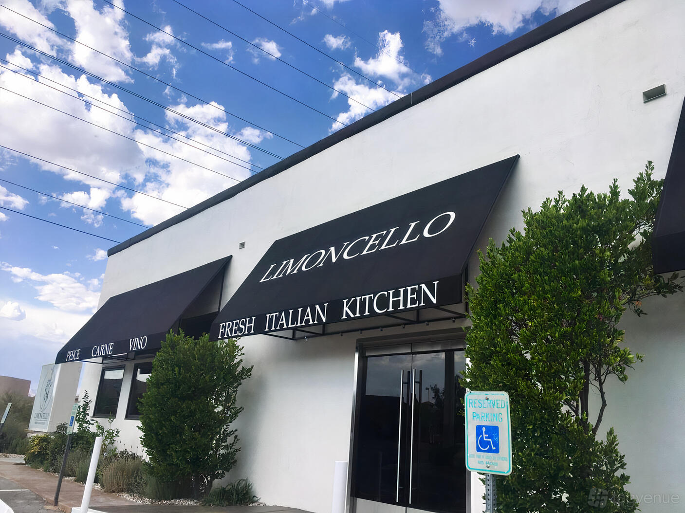 A restaurant facade with black awnings, white walls, and leafy green shrubs at Limoncello Fresh Italian Kitchen.