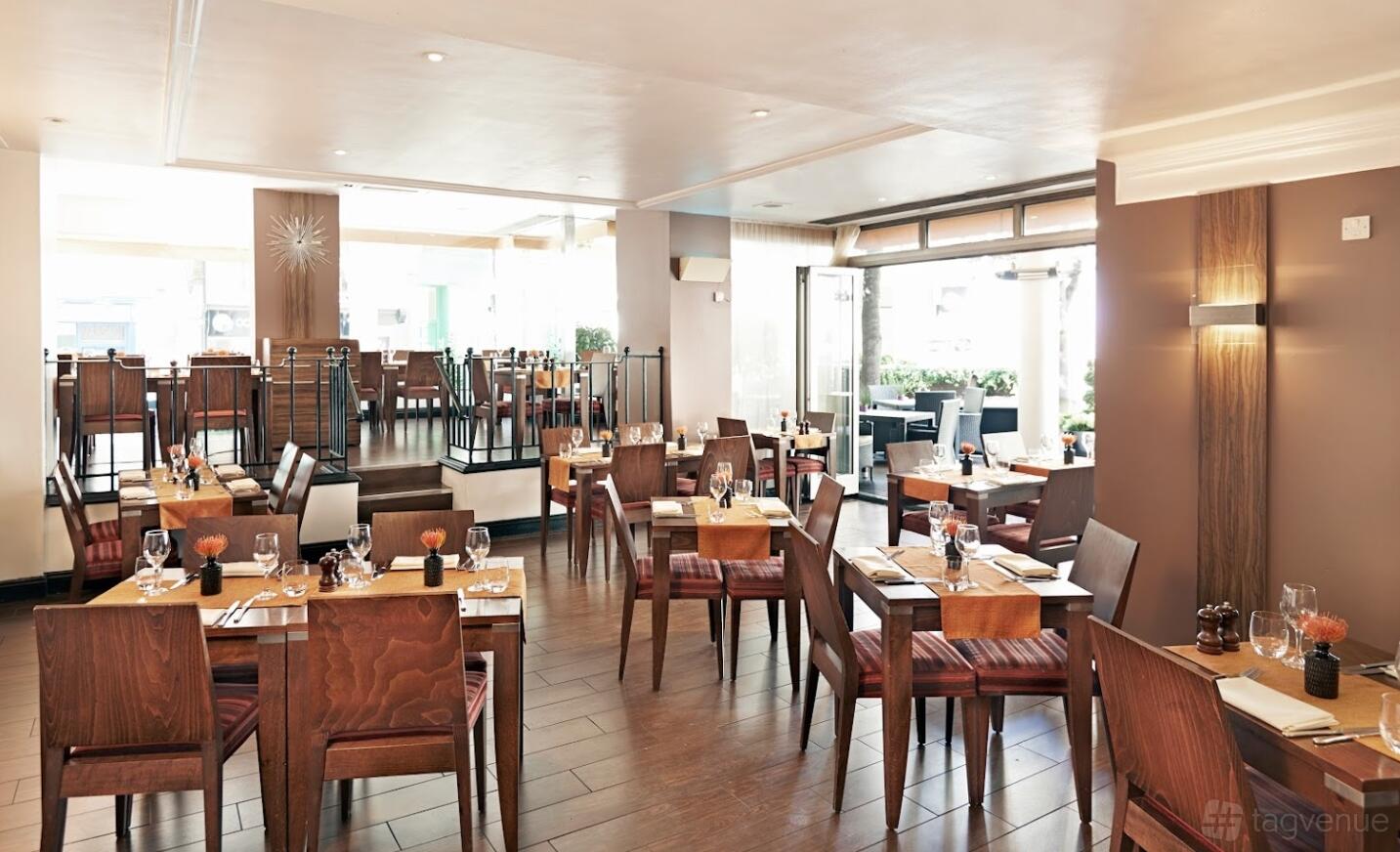 A restaurant with wooden tables, upholstered chairs, and floor-to-ceiling windows at Thistle Trafalgar Hotel.