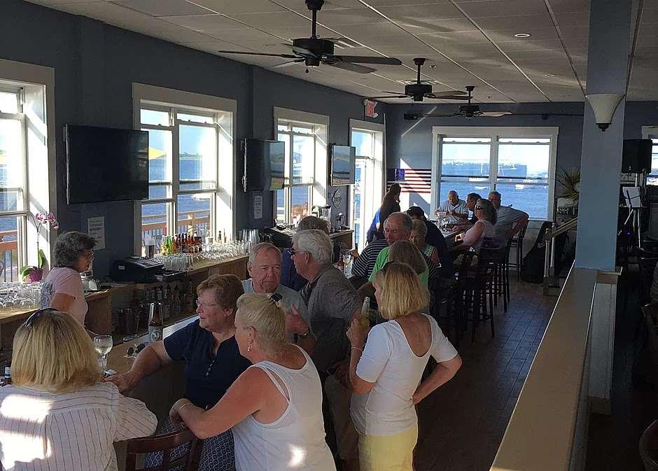 A restaurant with large windows, waterfront views, a long bar, and groups of people socializing at Bay House Breezy Point.