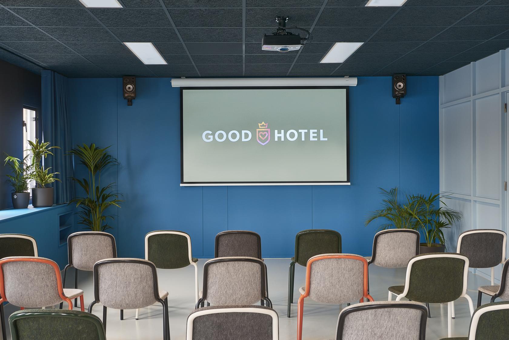A meeting room with a projector screen, rows of colorful chairs, and potted plants at Good Hotel London.