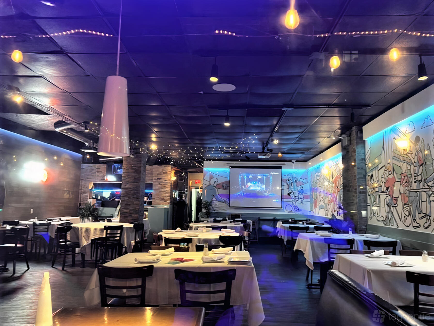 A restaurant dining area with white tablecloths, colorful mural walls, hanging lights, and a projector screen at New Campo Argentino.