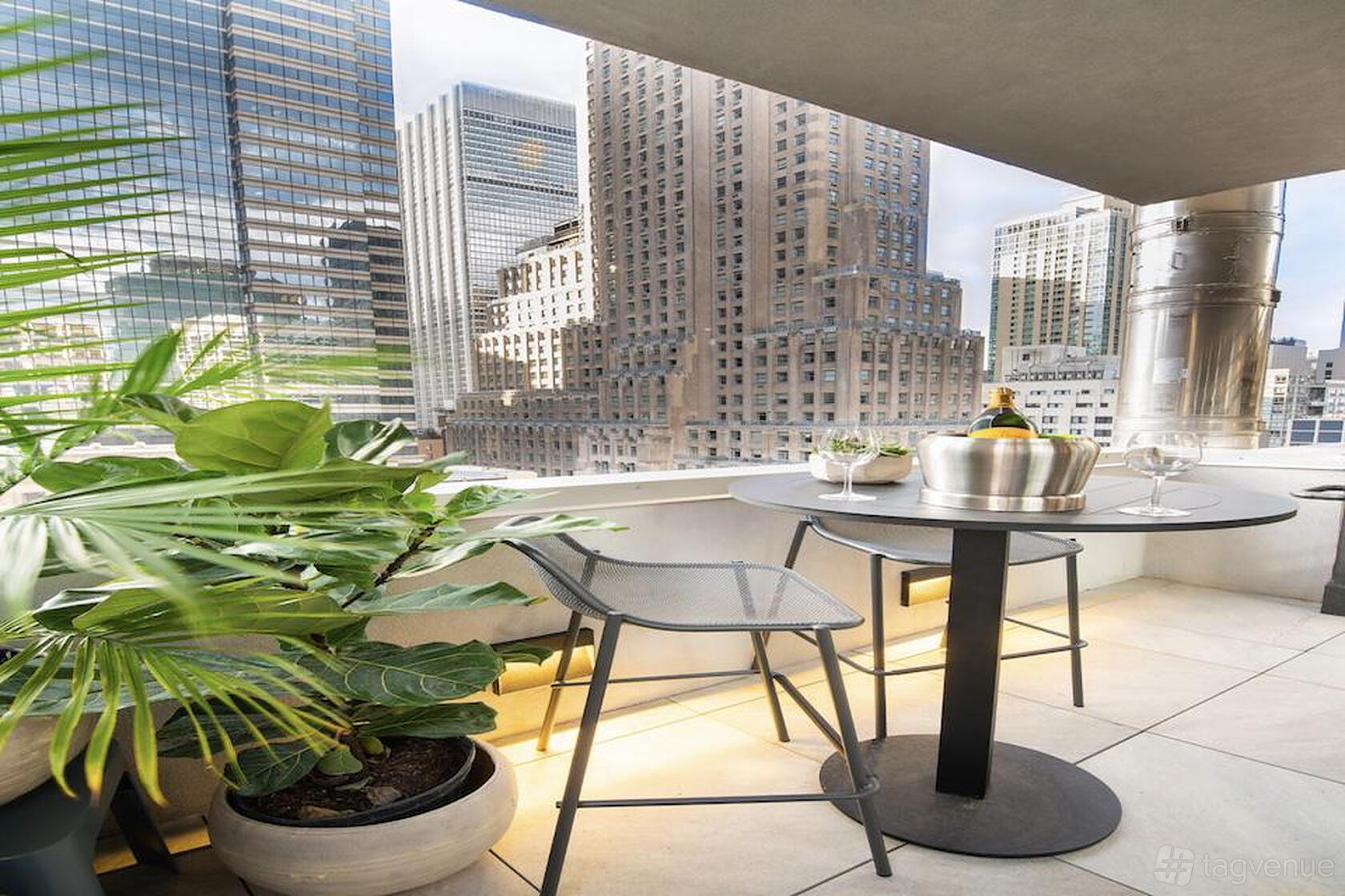 A terrace with potted plants, a small round table set for drinks, and city skyscraper views at Highwater Rooftop.