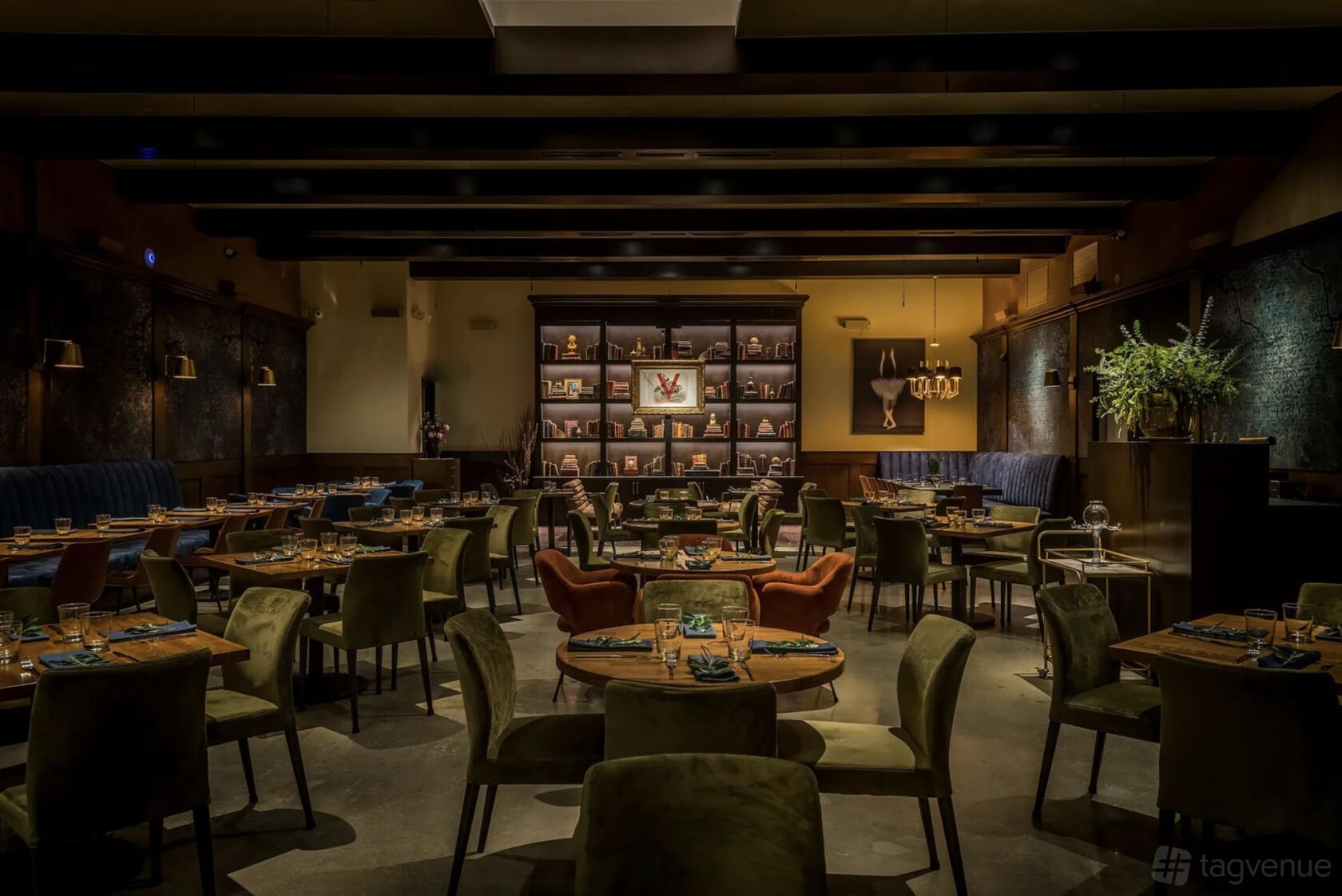 A restaurant with round wooden tables, velvet chairs, dim lighting, and a bookshelf wall at Verse.