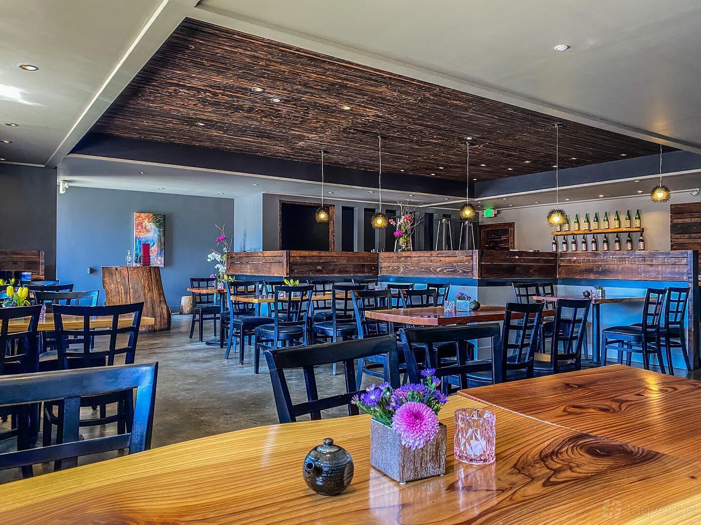 A restaurant dining room with wooden tables, black chairs, pendant lighting, and a display of sake bottles at AKEMI Japanese Restaurant.