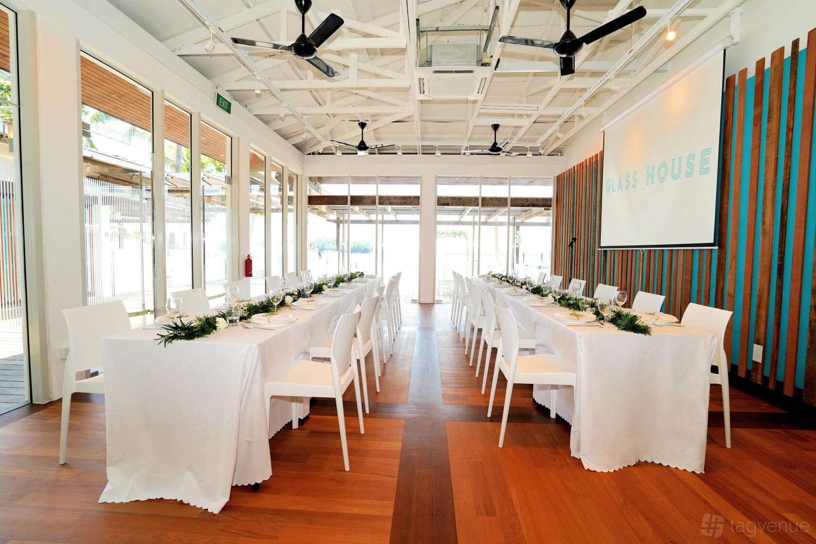 A meeting centre with floor-to-ceiling windows, ceiling fans, and long banquet tables with white linens at Glass House.