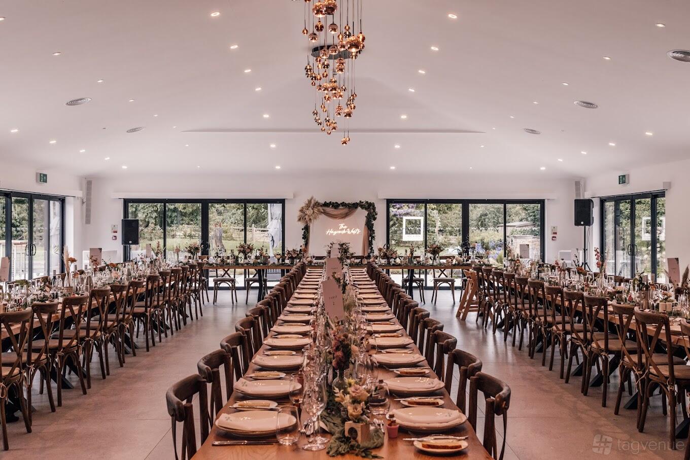 A barn with long banquet tables, wooden chairs, floral centerpieces, and glass doors at Chapel House Estate.