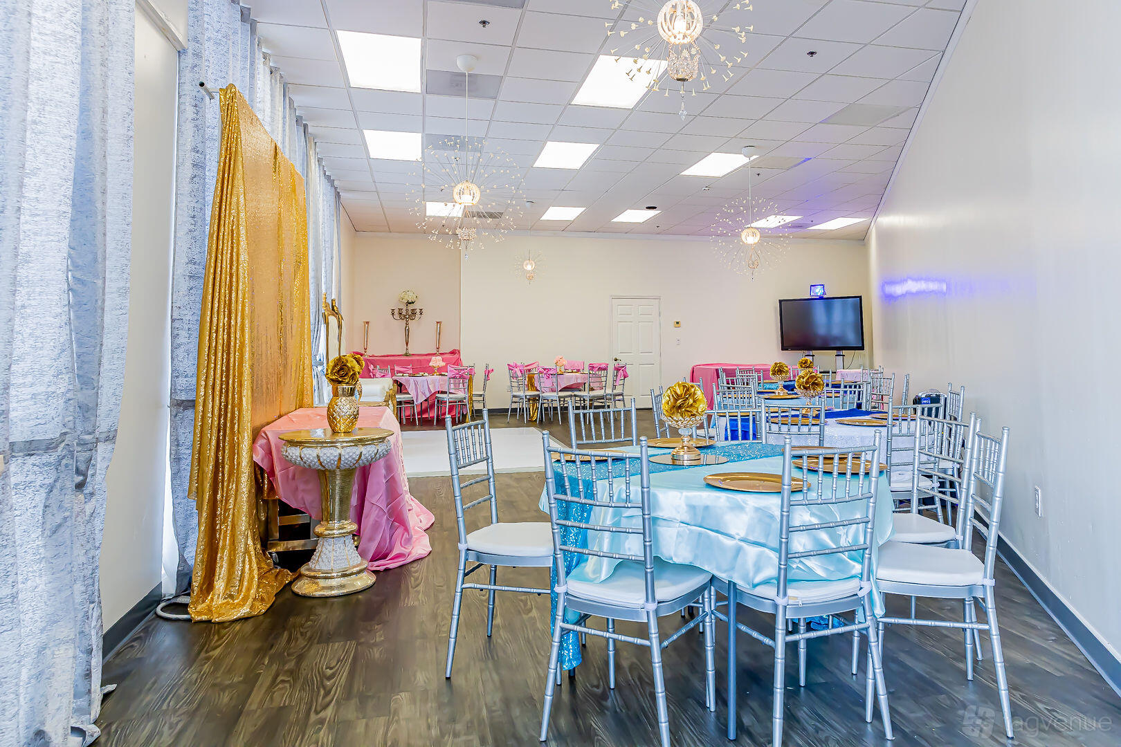 An event venue with round tables covered in light blue linens, gold centerpieces, and silver chairs at Hillcroft Venue.